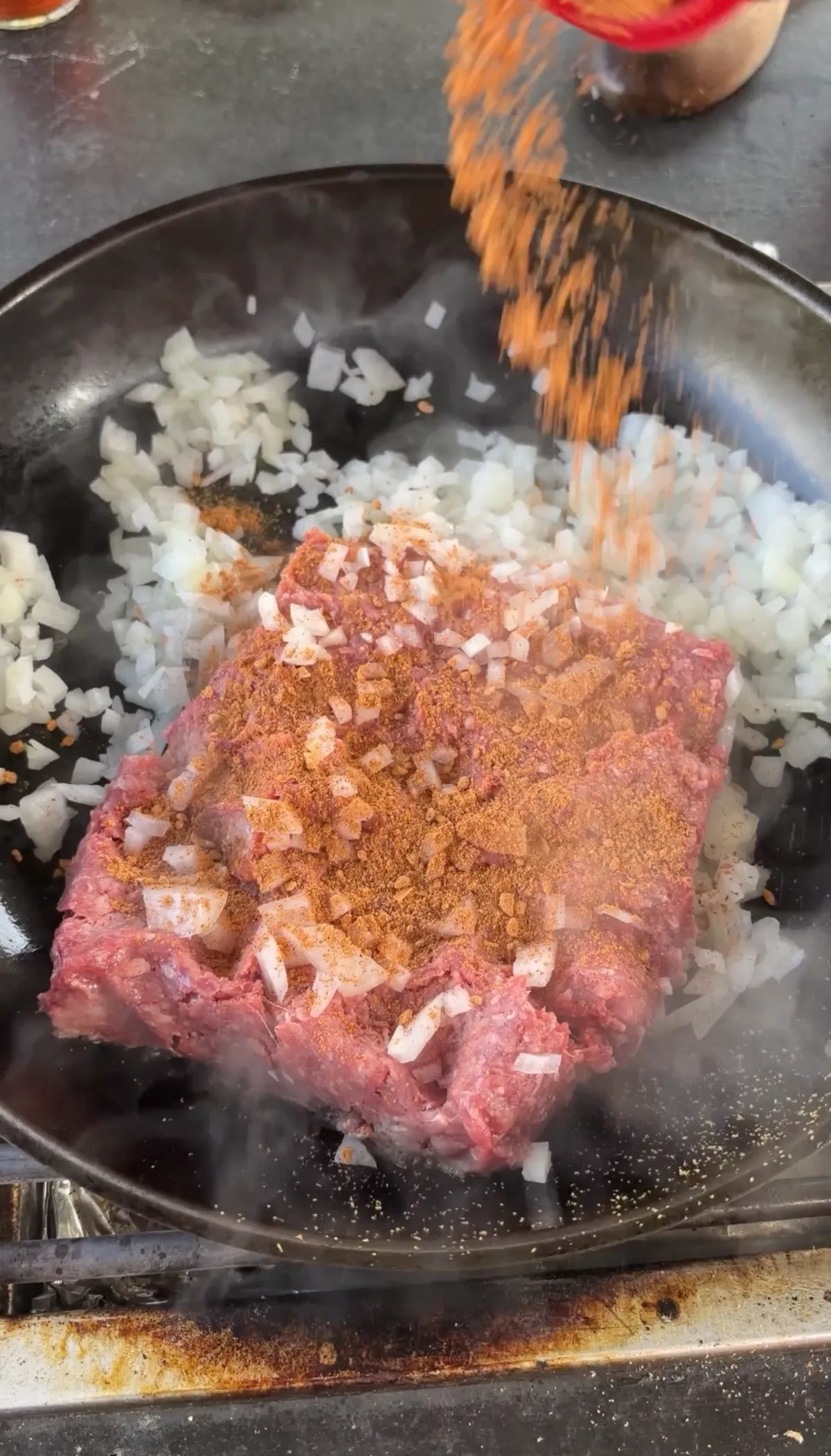Taco seasoning being poured over 85/15 ground beef and diced onion cooking in a hot skillet