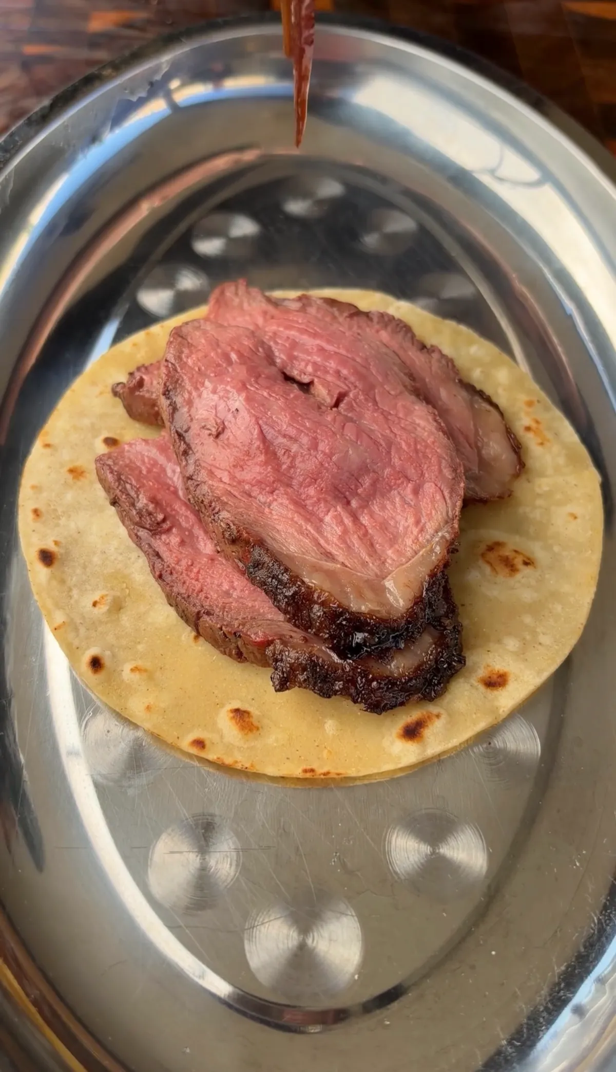 Thinly sliced rotisserie picanha on a warm corn tortilla served on a silver tray with salsa drizzling from above