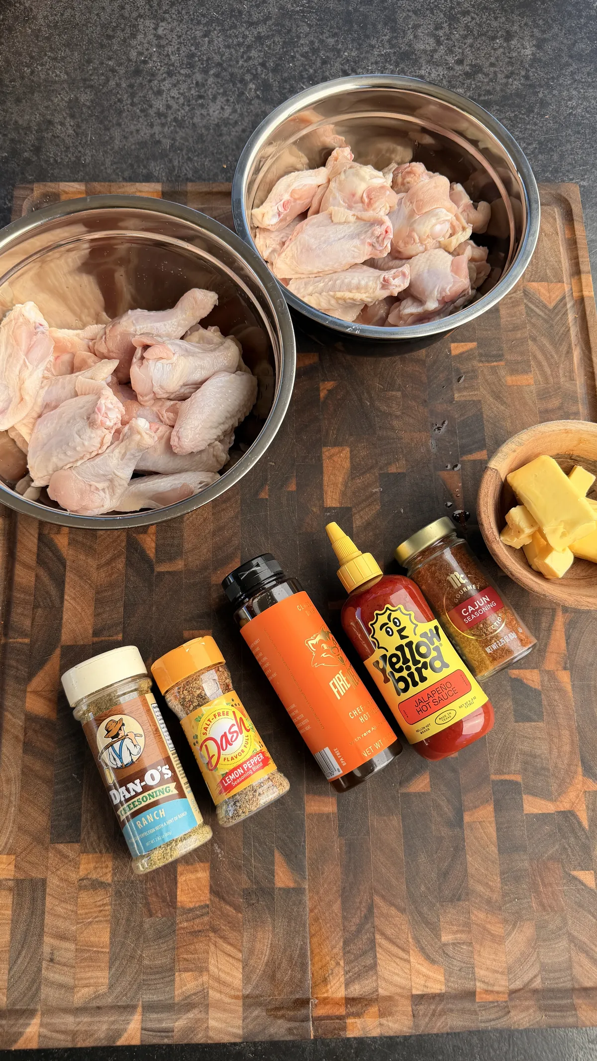 Raw chicken wings in two bowls with Dan-O's Ranch, lemon pepper, Cajun seasoning, jalapeño sauce, and butter on a cutting board