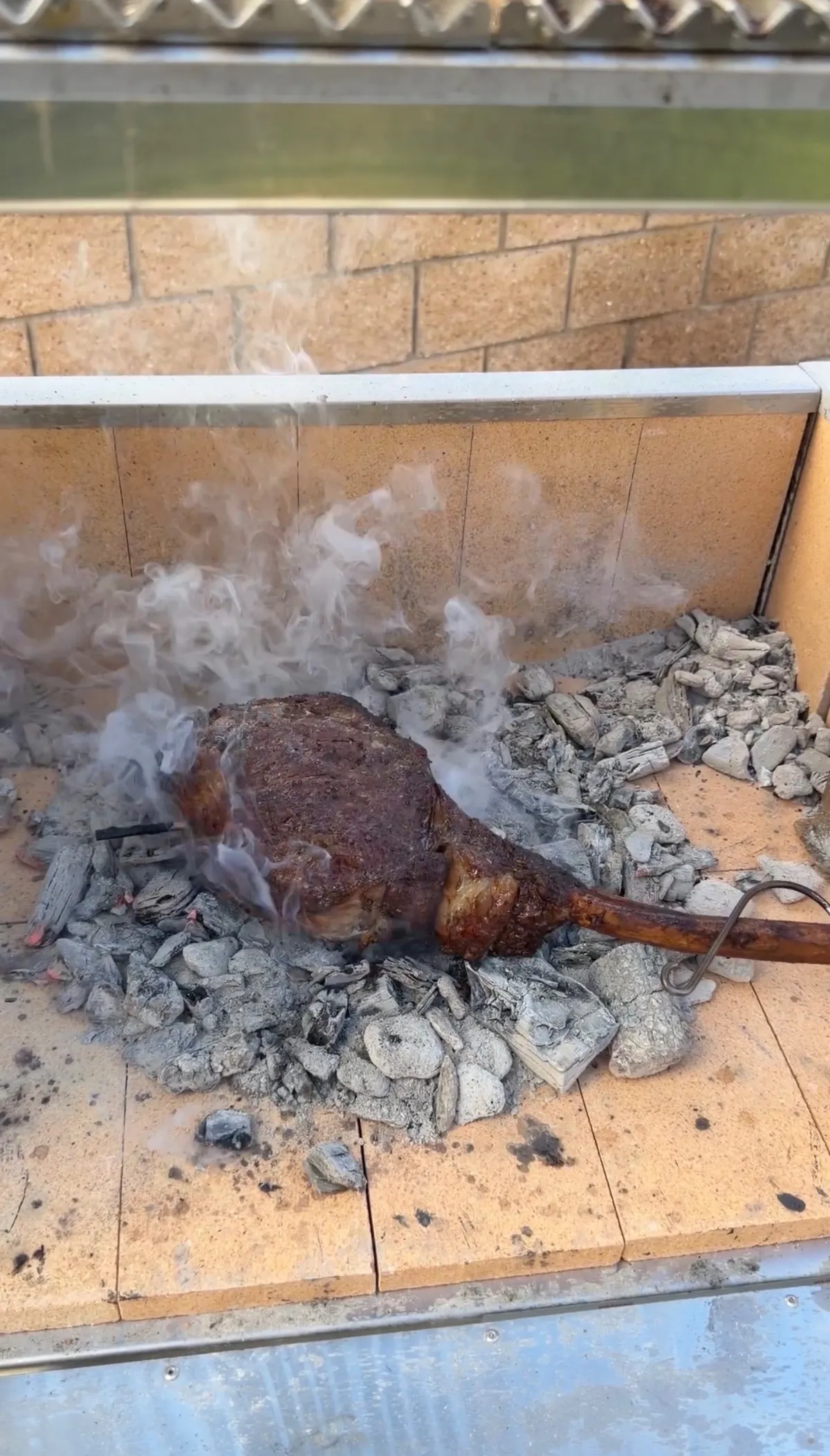 Tomahawk steak caveman-searing directly on hot lump charcoal with smoke rising in a brick firebox
