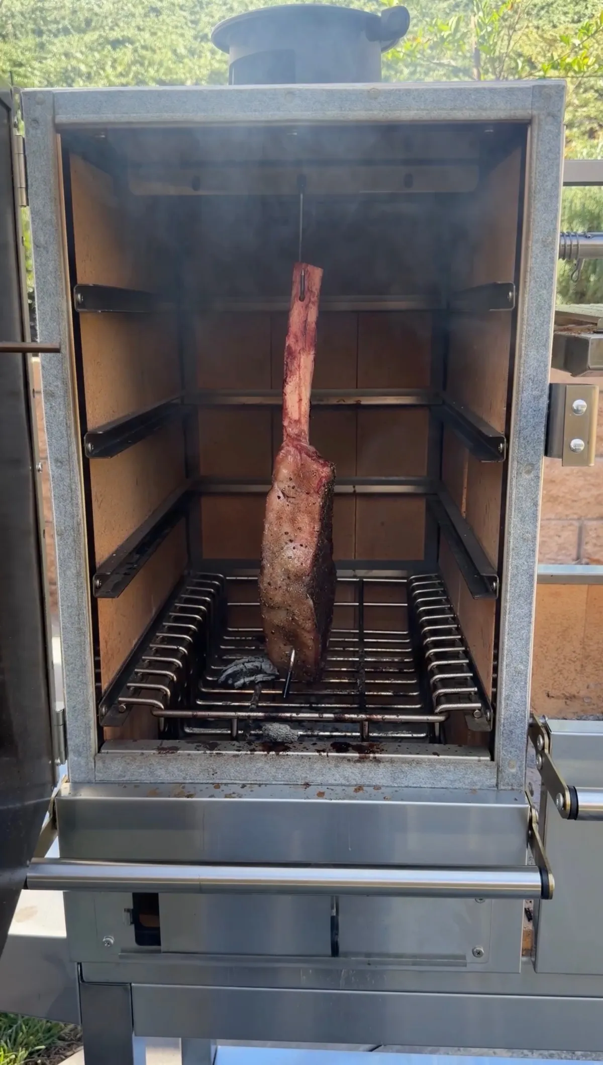 Tomahawk steak hanging by hook inside smoker over grates, surrounded by Applewood smoke during low and slow cook