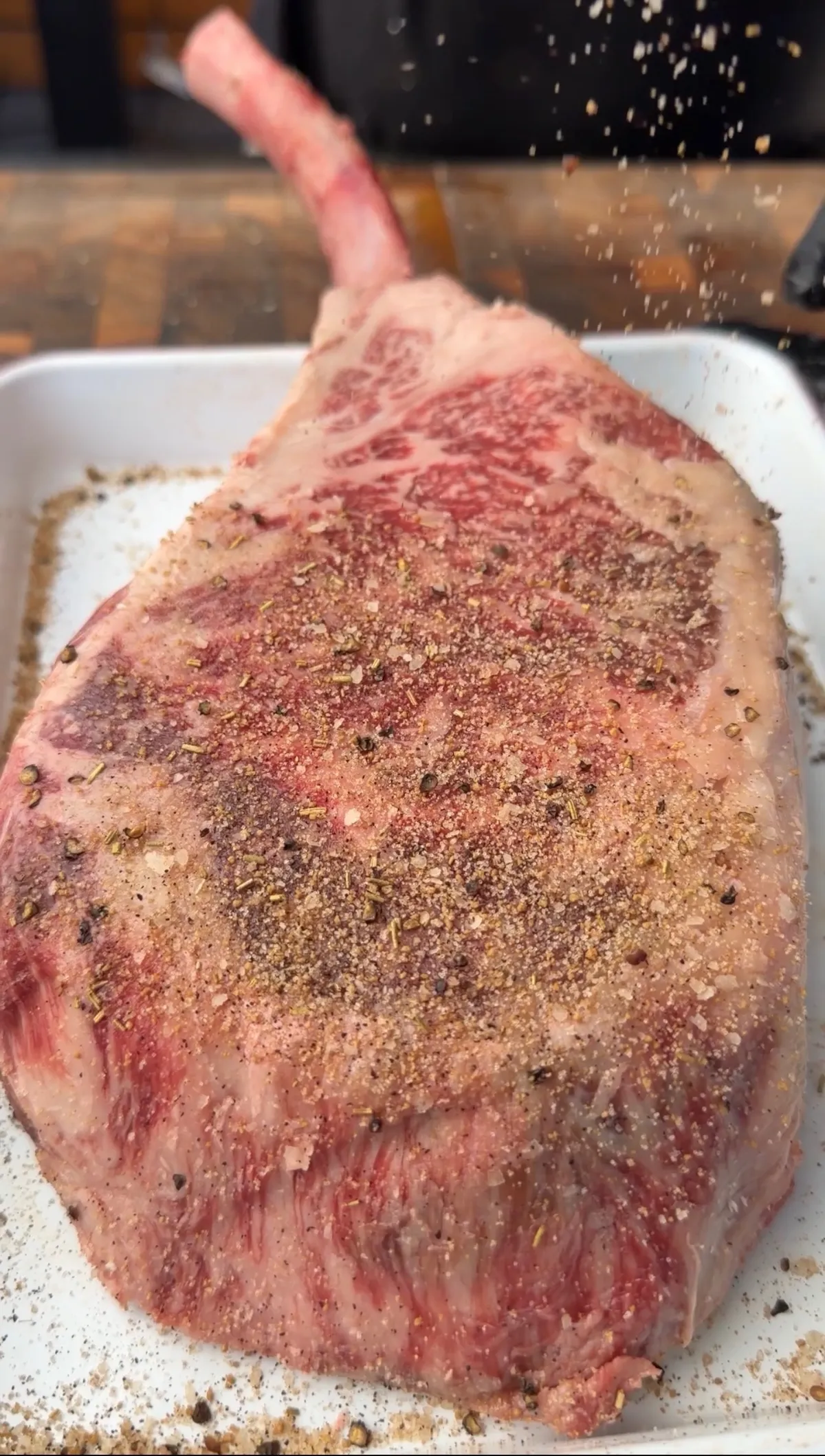 Raw tomahawk steak being seasoned with kosher salt, coarse black pepper, and garlic powder in a white tray