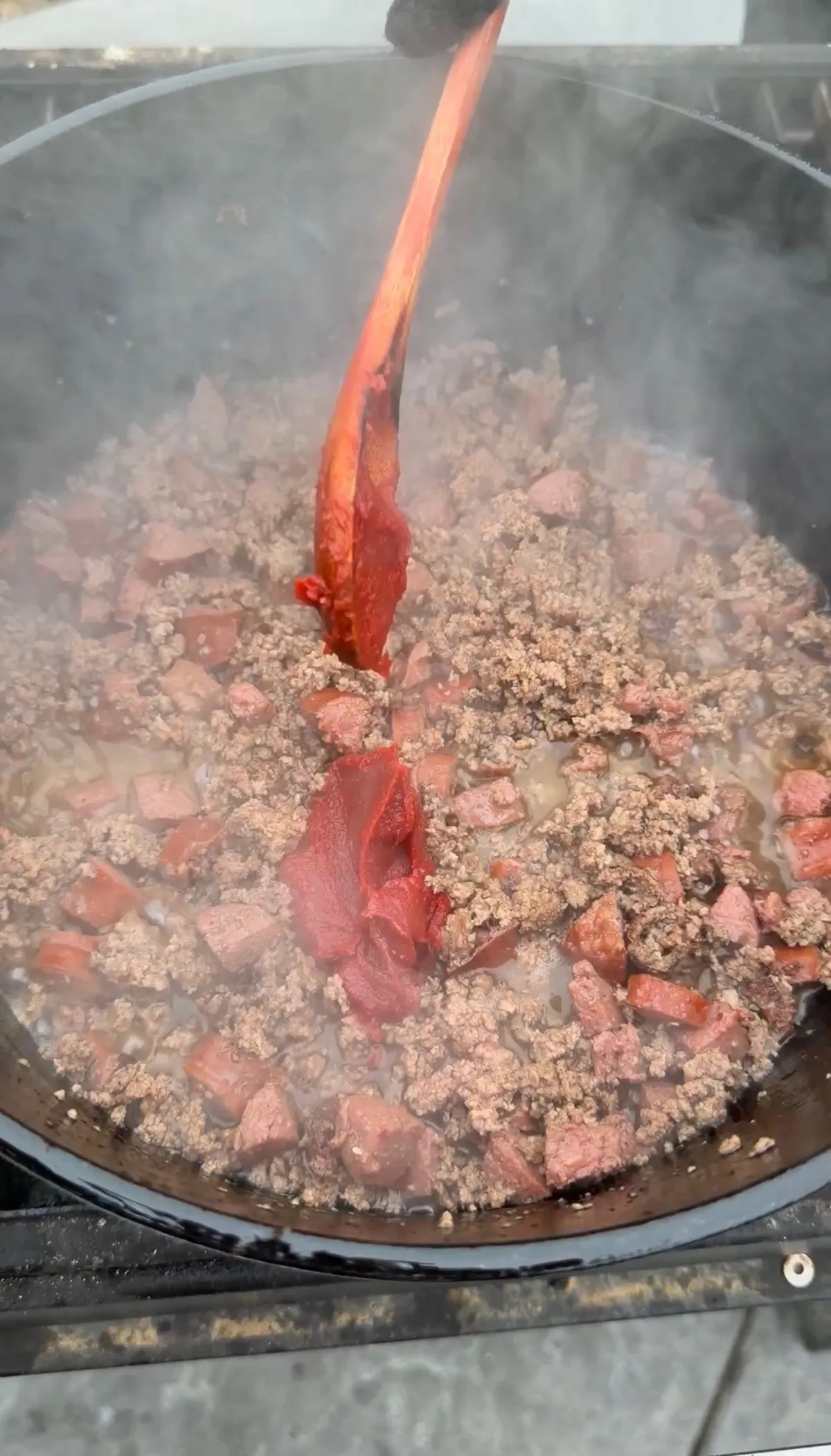 Tomato paste being stirred into browned ground chuck and kielbasa in a large Dutch oven, steaming hot
