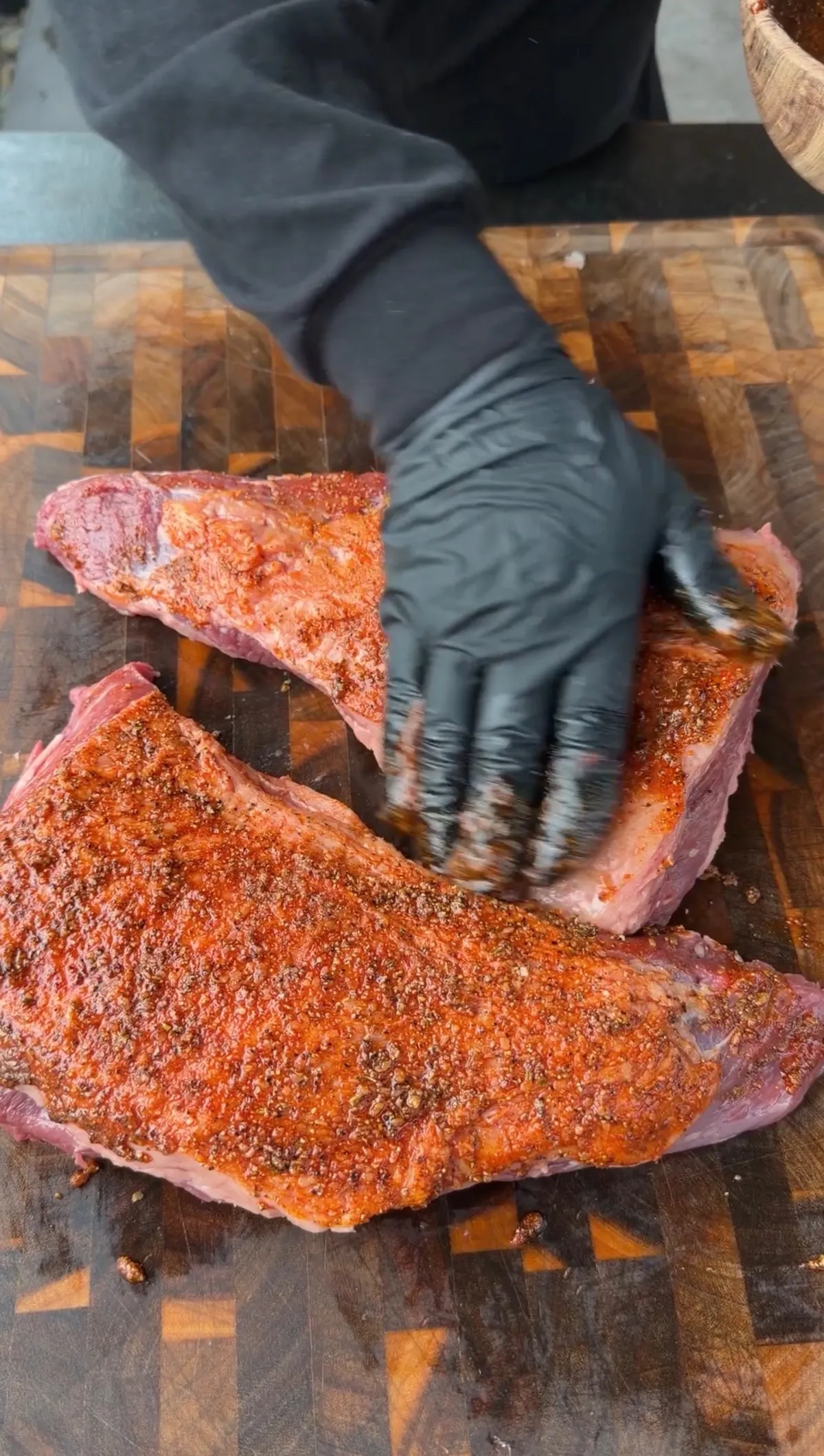Gloved hands rubbing seasoning mud onto two raw tri-tip roasts on a wood cutting board