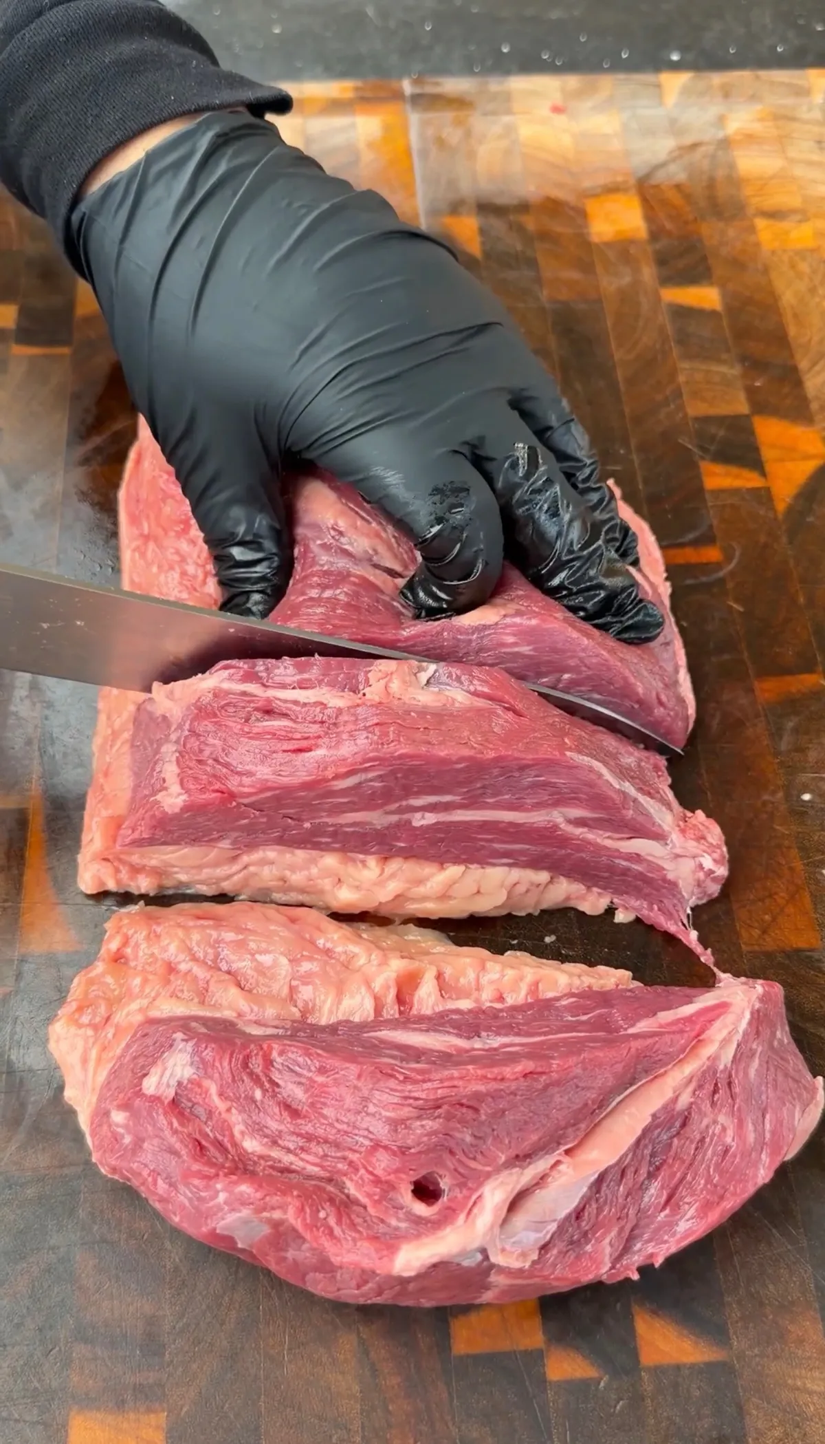 Gloved hands slicing whole tri-tip into 1-inch-thick steaks on a wood cutting board for taco skewers