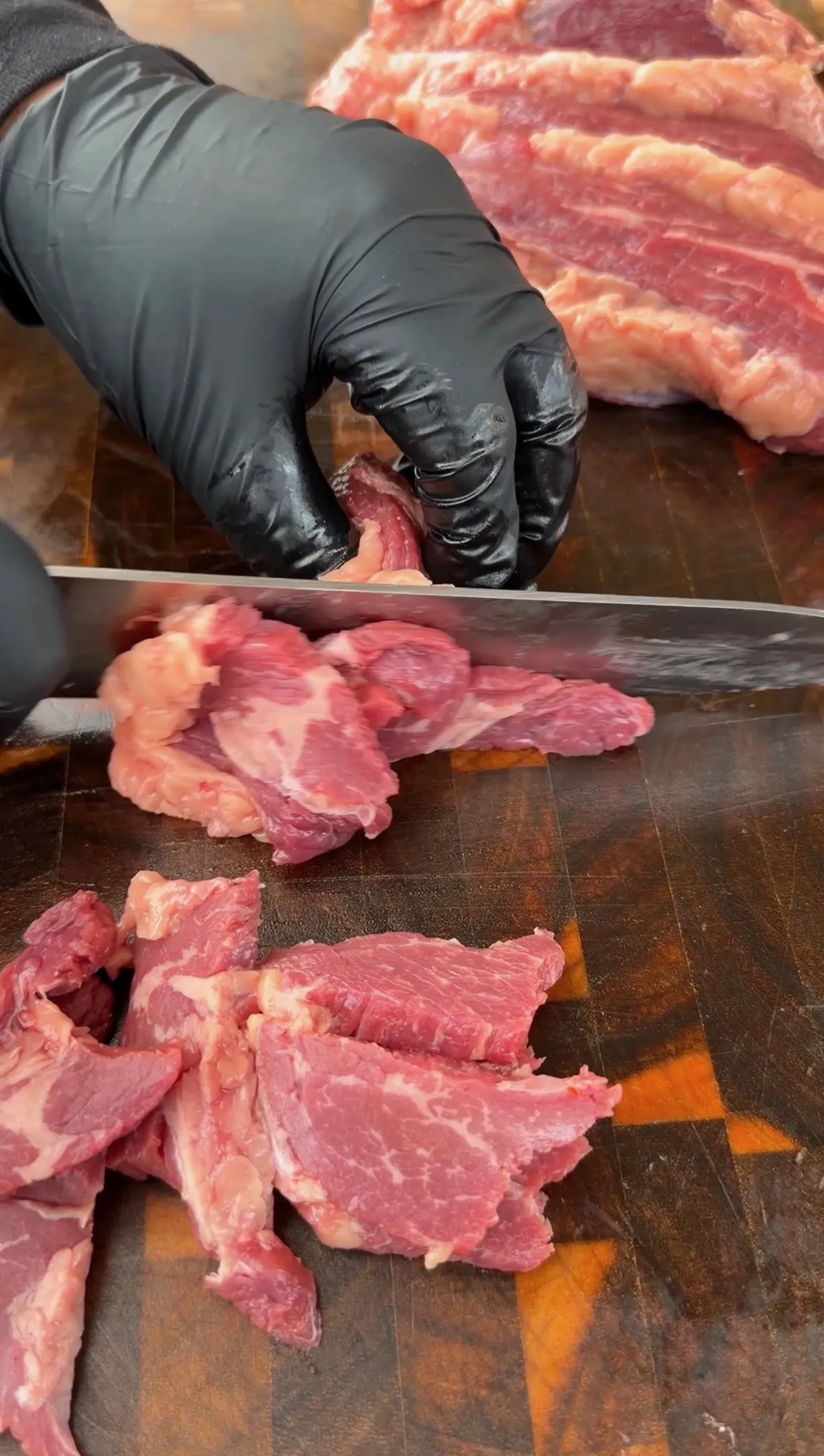 Gloved hands slicing tri-tip thin against the grain on a wooden cutting board for taco skewers