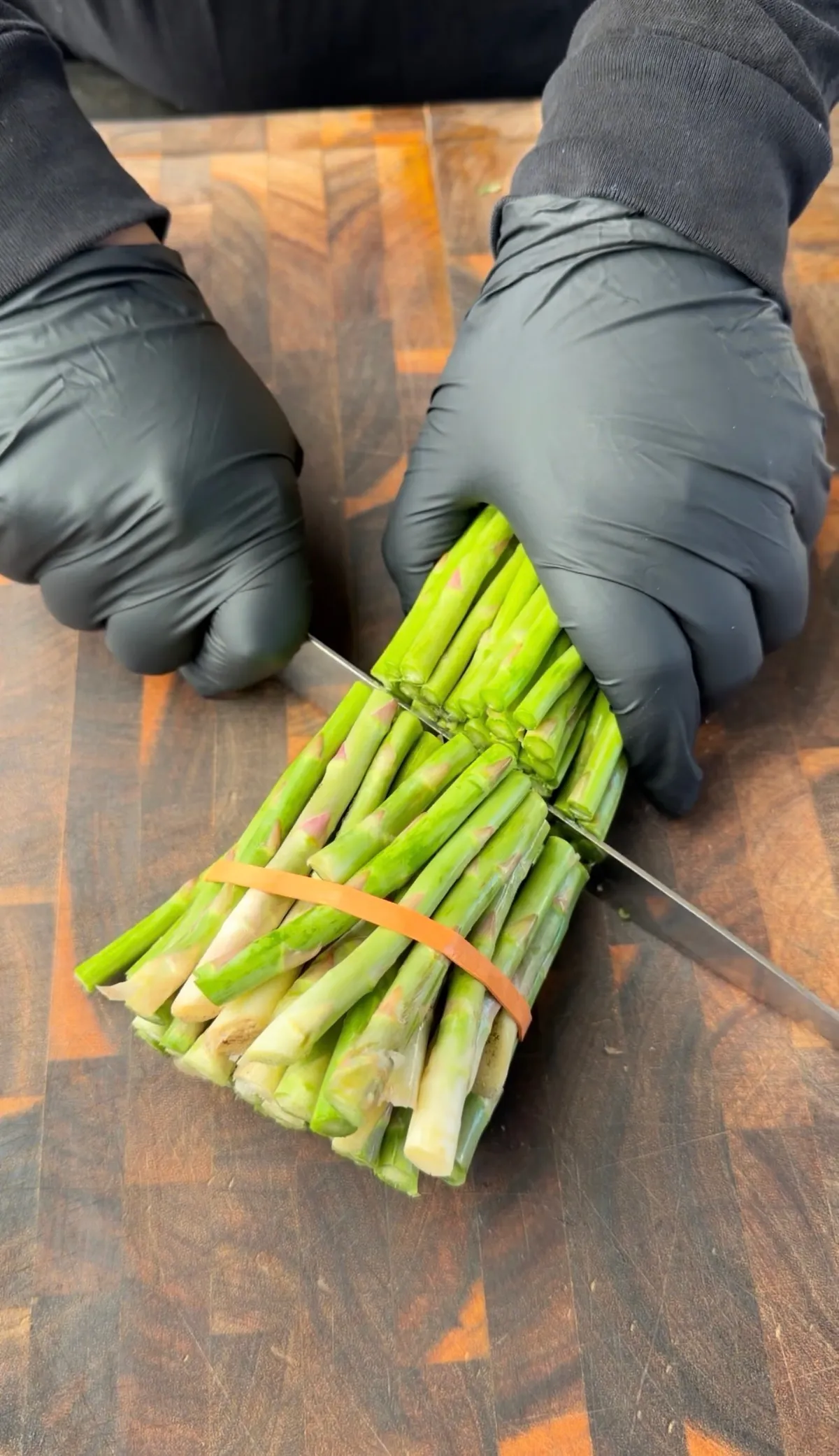 Gloved hands trimming woody ends off a bound bundle of asparagus with a knife on a wooden cutting board
