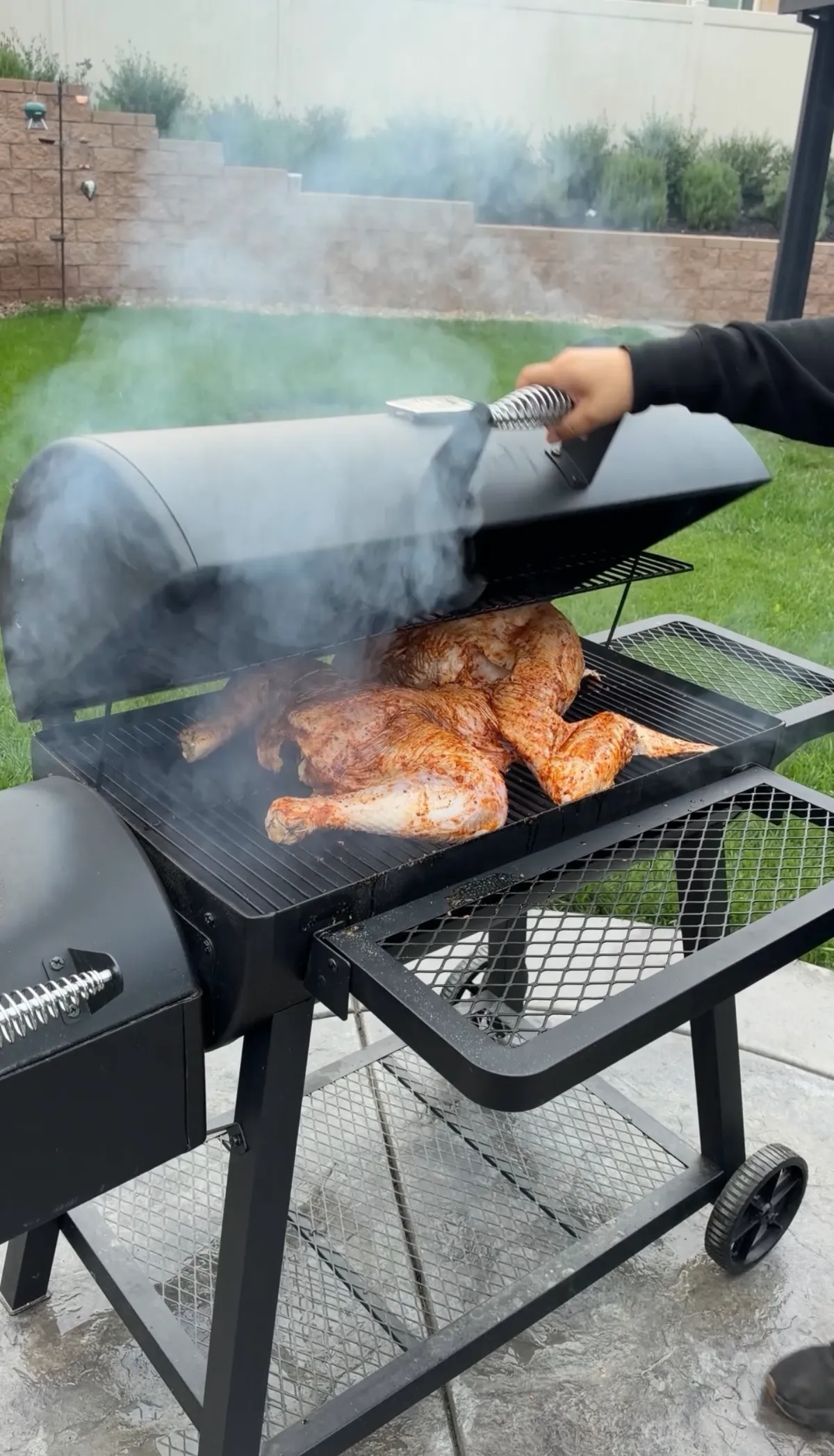 Hand lifting smoker lid to reveal Cajun-seasoned frog-style turkey smoking on grill grates with billowing smoke