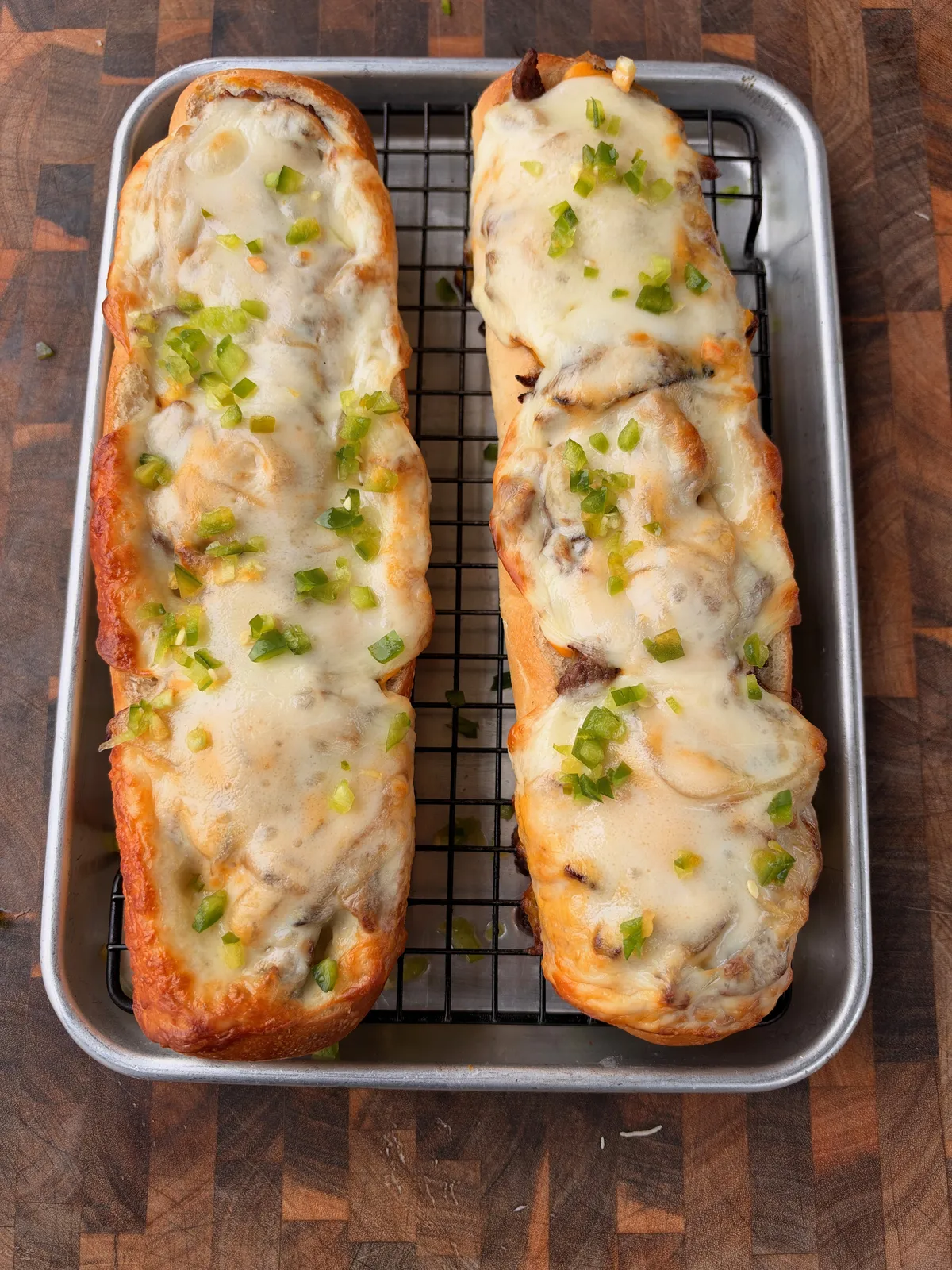 Two cheesesteak stuffed French baguette boats with melted provolone and diced jalapeños on a wire rack baking pan