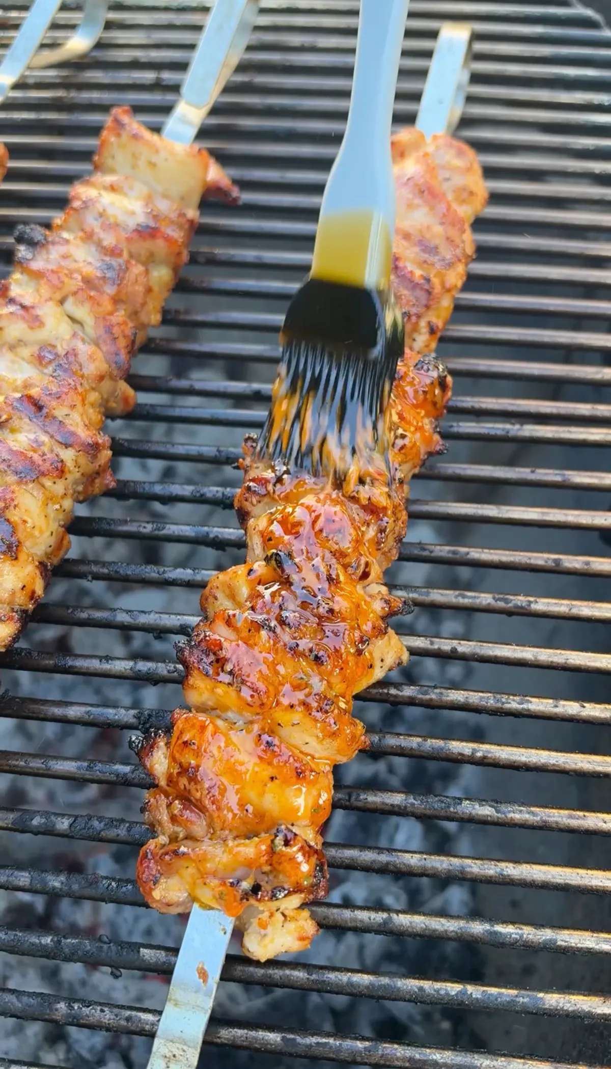 Silicone basting brush applying cowboy butter onto grilled chicken thigh skewers over charcoal grill grates