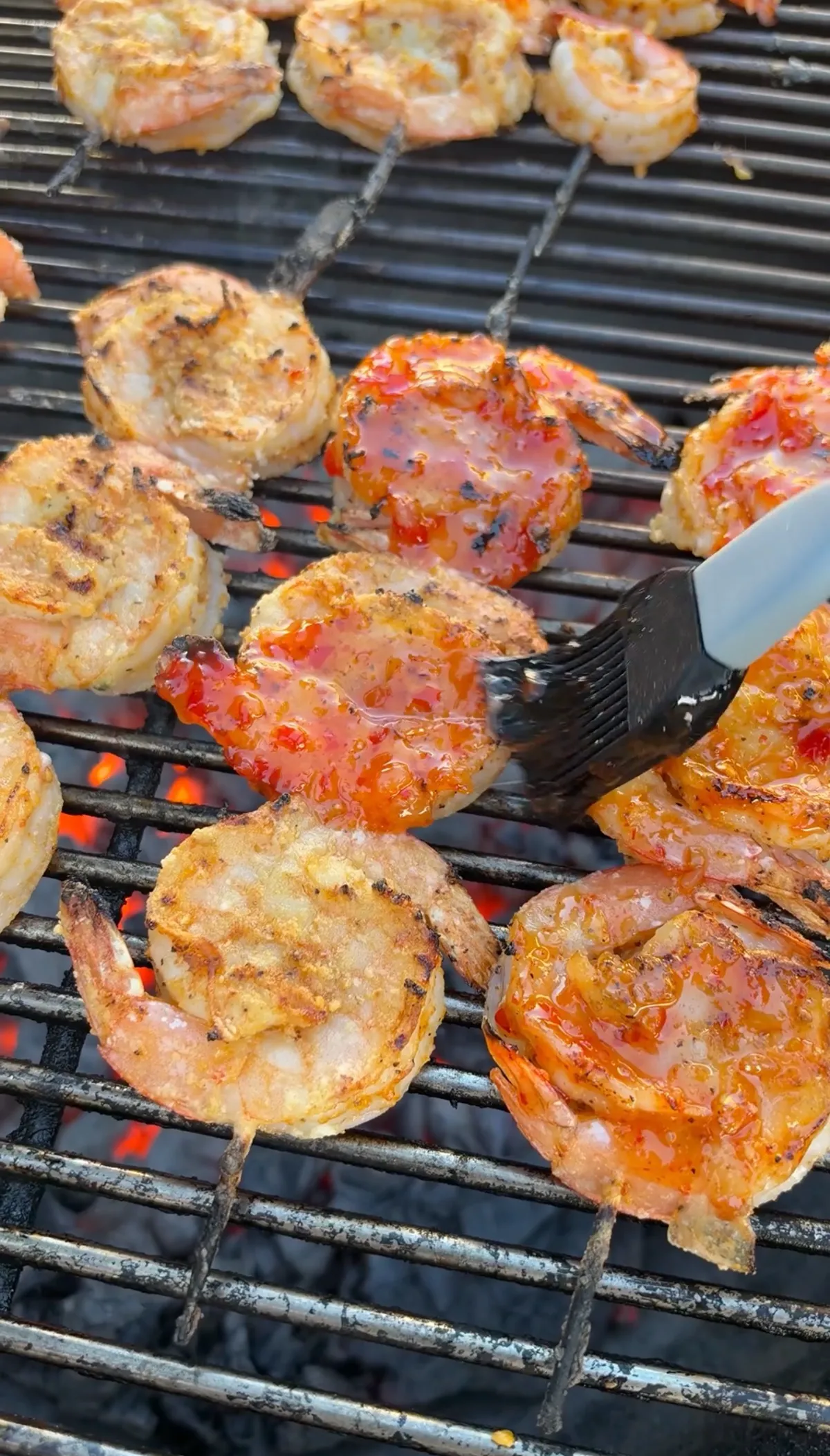 A silicone basting brush applying sticky firecracker glaze to grilled shrimp skewers over hot charcoal grill grates