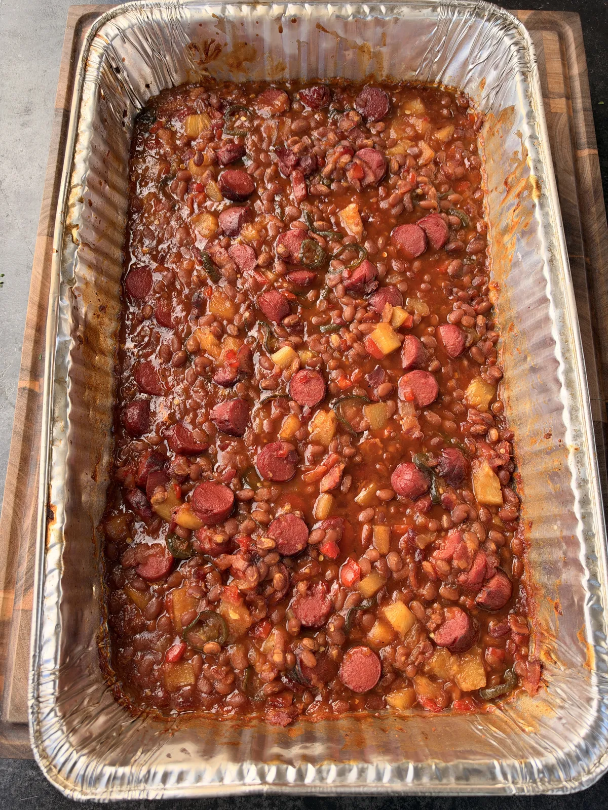 Smoked BBQ baked beans with sliced hot dogs, pineapple tidbits, and jalapeños in a large foil pan