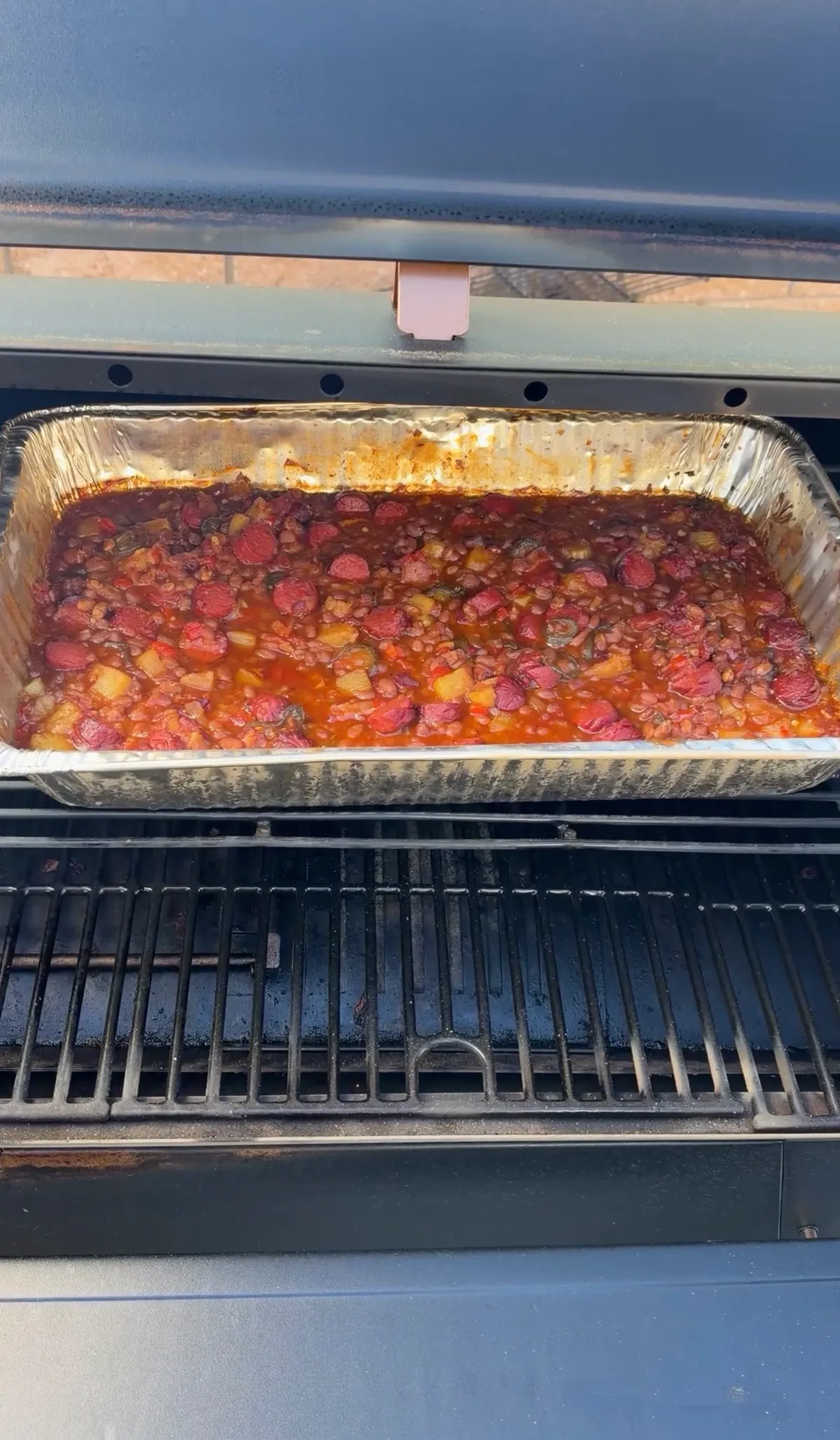 Smoked BBQ baked beans with hot dog rounds and pineapple in a foil pan on pellet smoker grates at 250°F