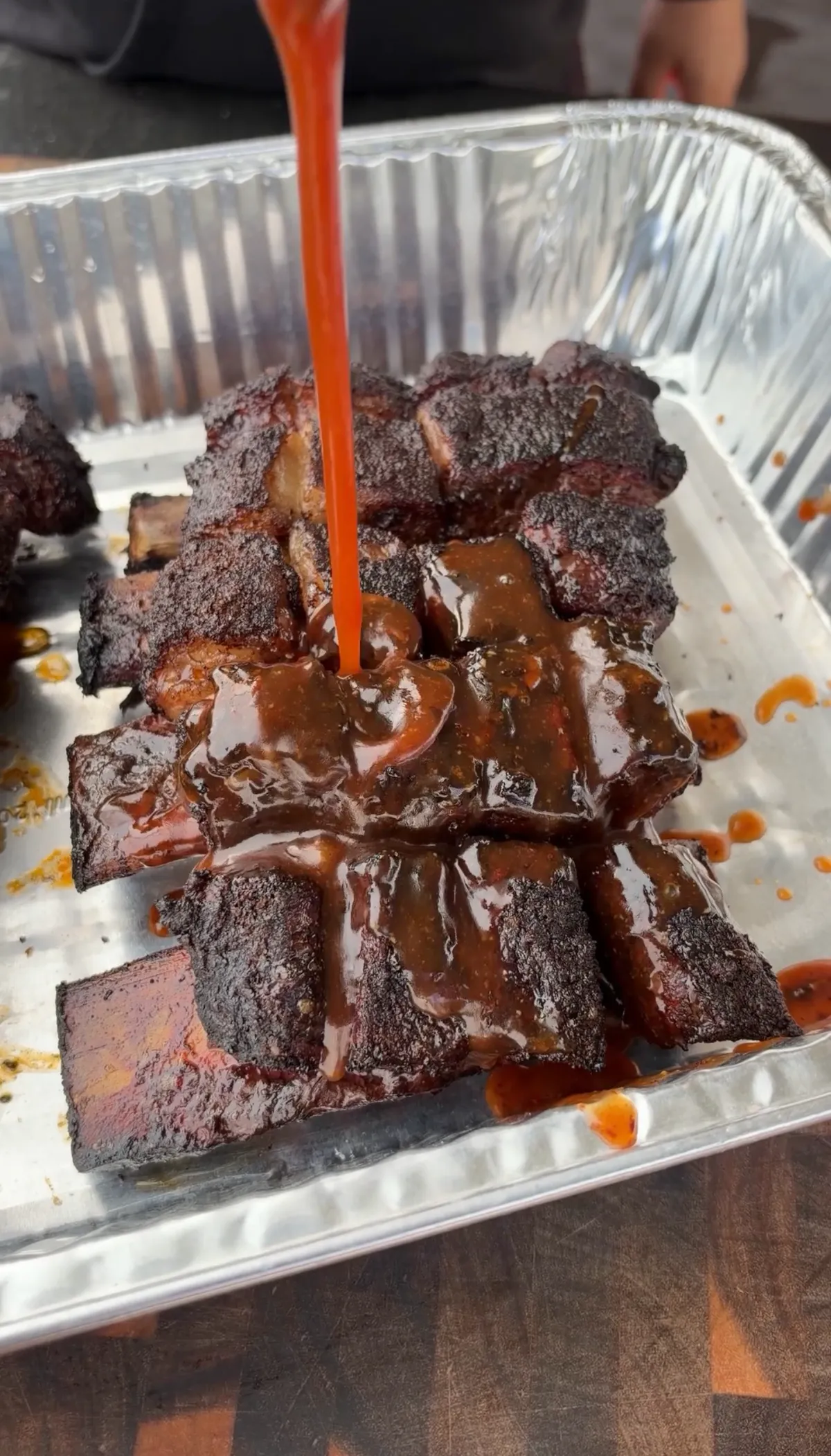 Dark BBQ sauce being poured in a stream over smoky, caramelized beef short rib burnt ends in an aluminum pan