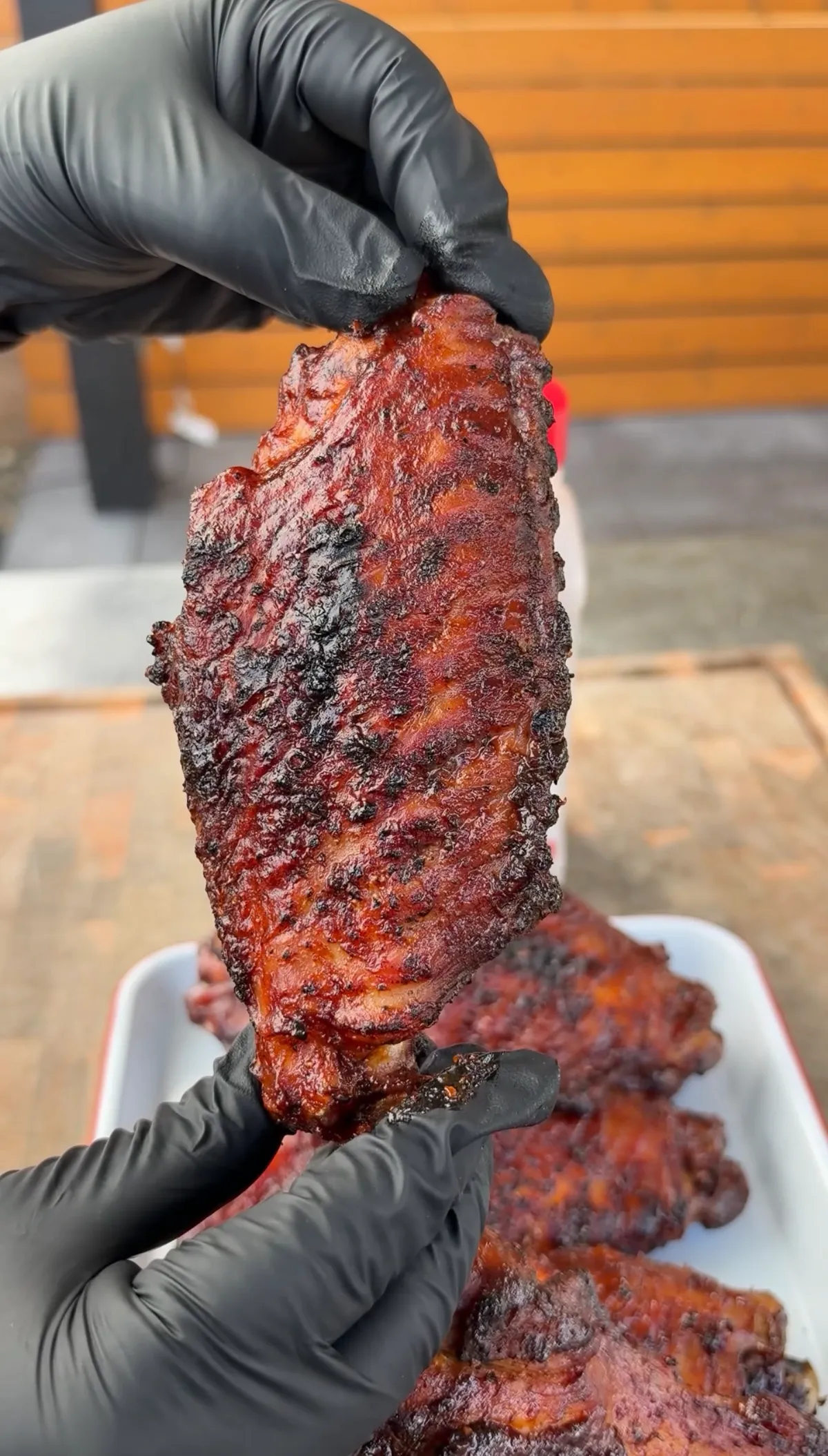 Gloved hands holding a caramelized BBQ-glazed grilled turkey wing flat above a white tray of wings