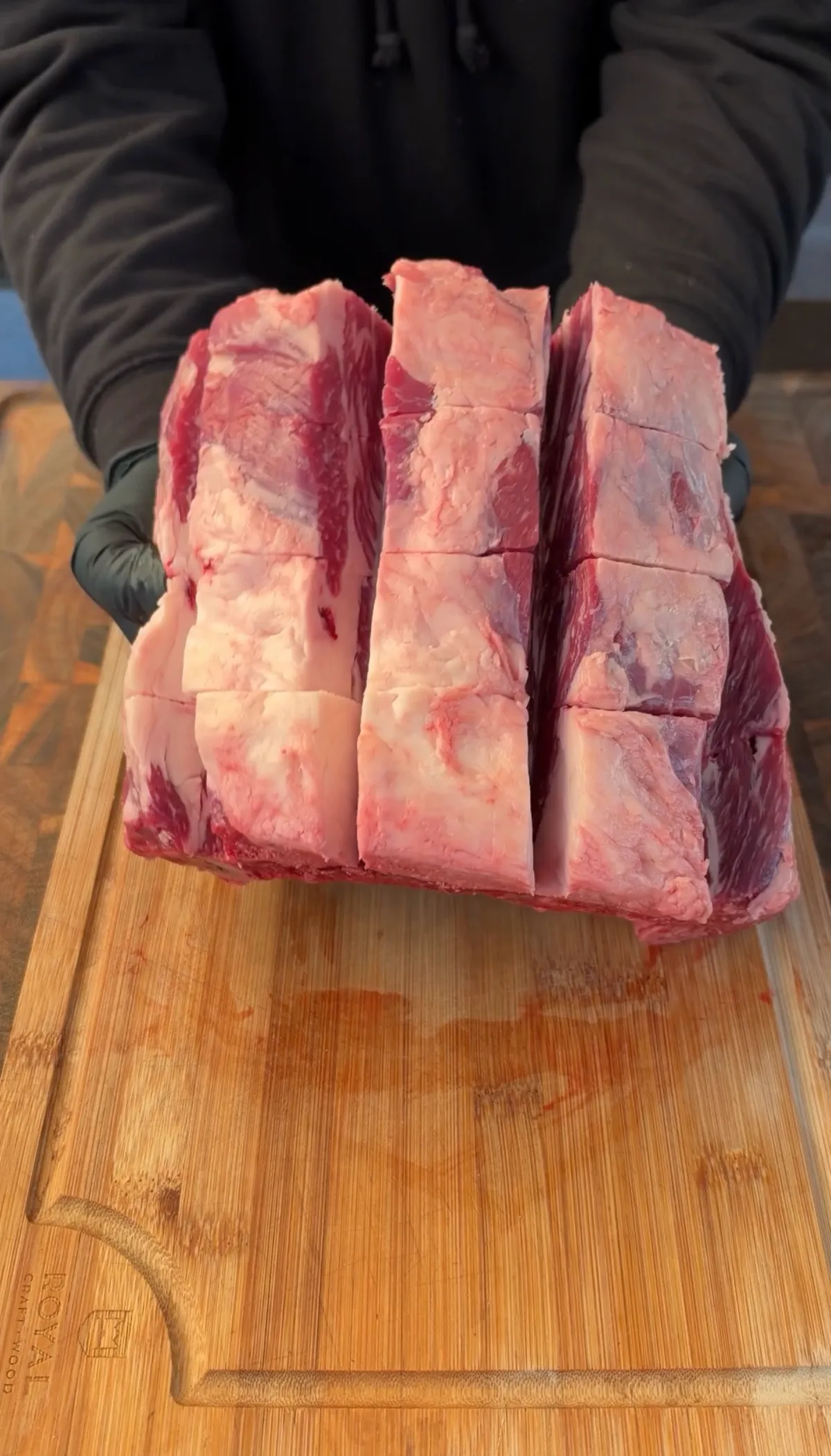 Hands in black gloves holding a rack of raw beef short ribs sliced into cubes on a bamboo cutting board