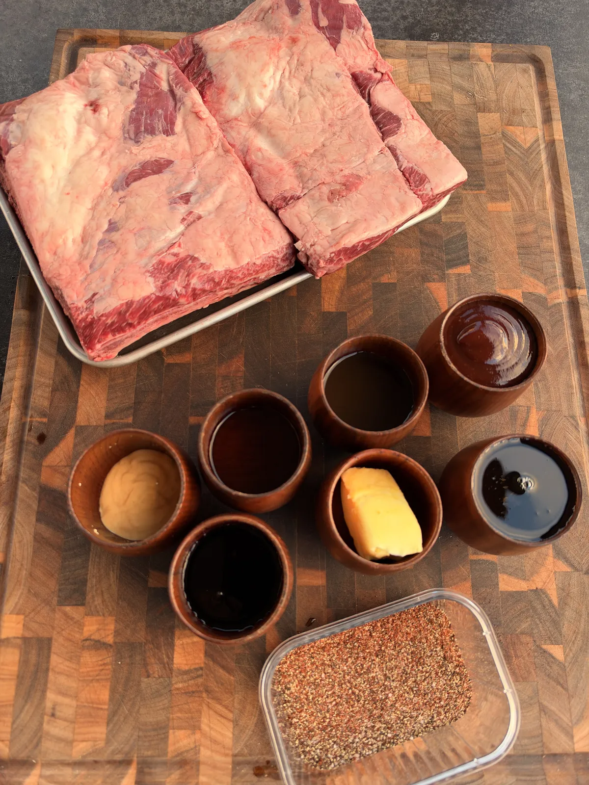 Raw beef short ribs with small bowls of sauces, mustard, butter, spice rub ingredients on a wooden cutting board