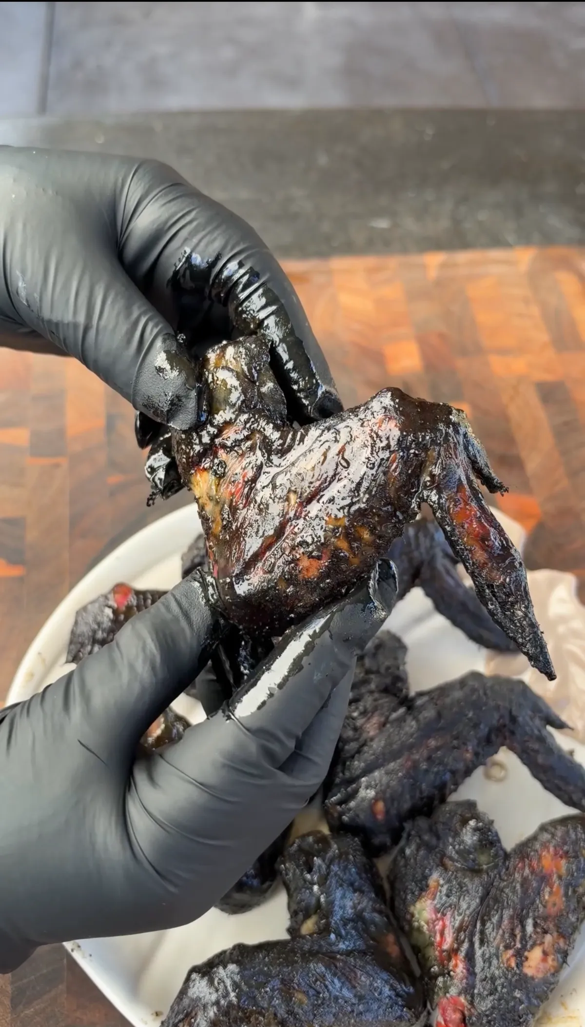 Hands in black gloves holding a grilled black chicken bat wing with deep charred marinade over a plate of wings