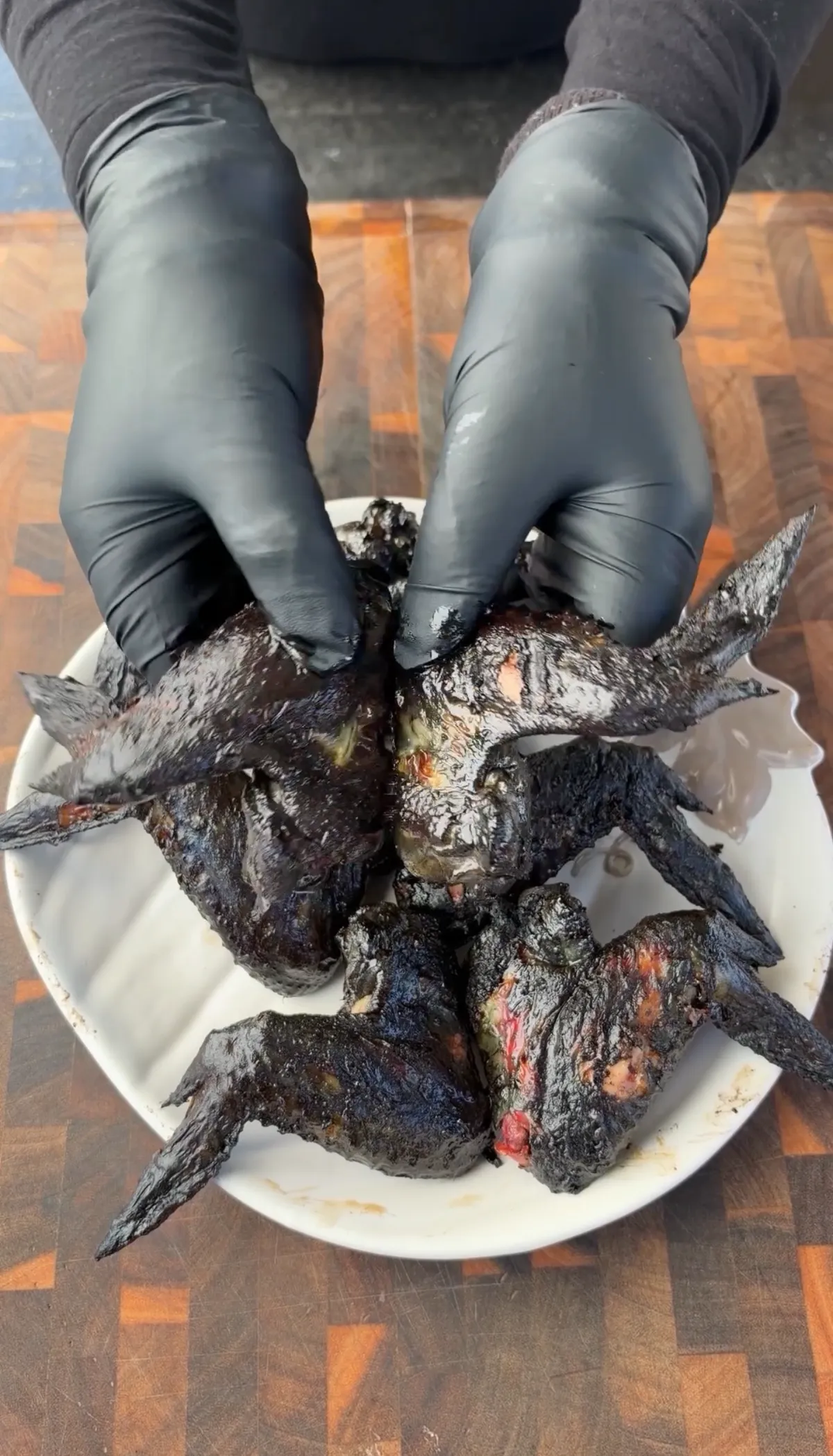 Grilled black chicken bat wings with deep black marinade arranged on white plate, held by black gloved hands