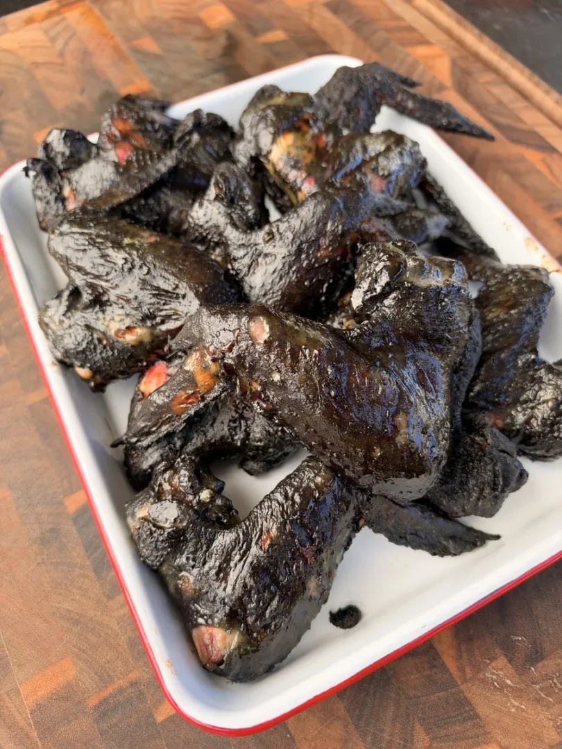 Grilled black chicken bat wings coated in soy-honey-chili marinade piled in a white enamel tray on a wood cutting board