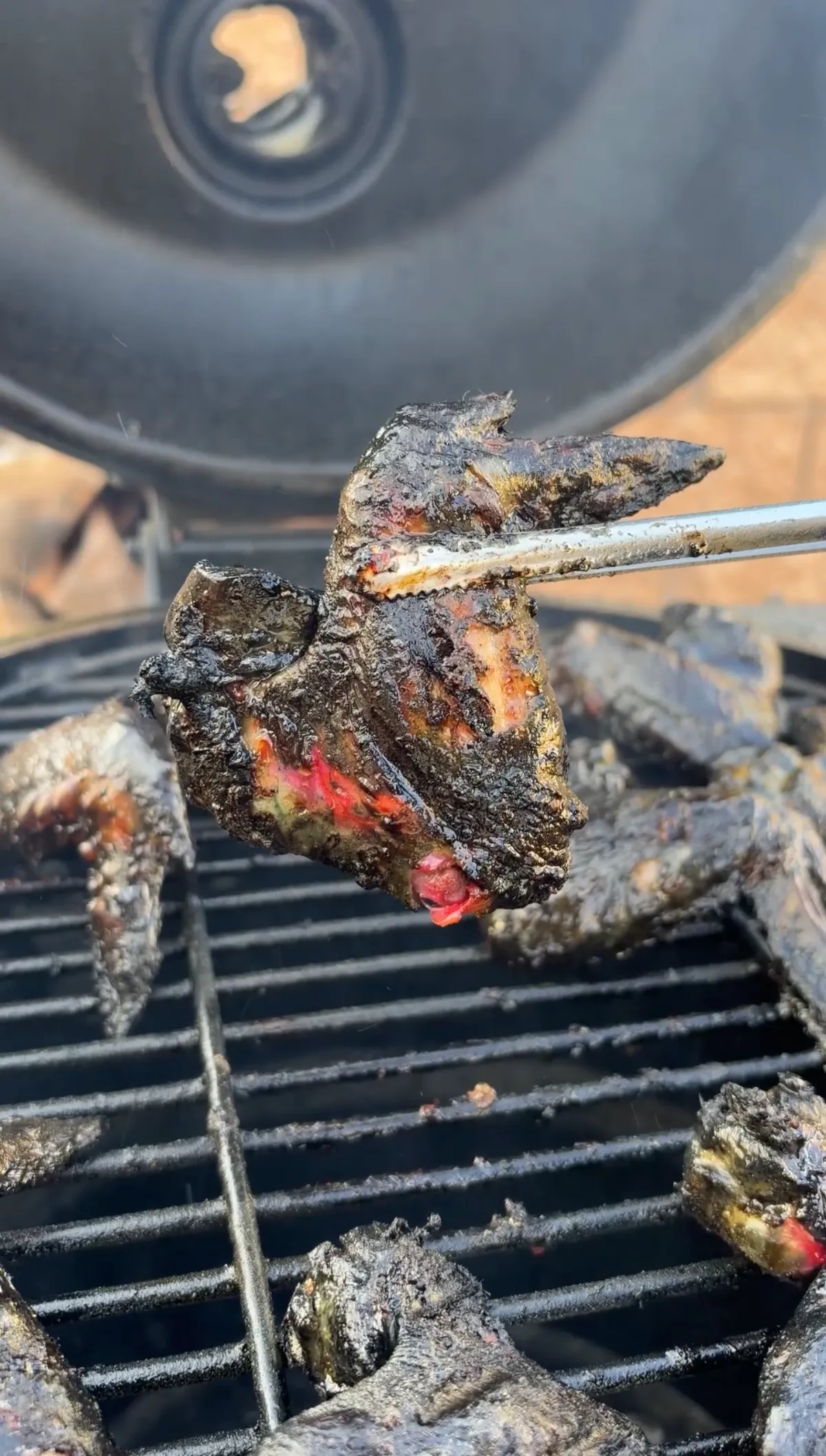 Black marinated chicken wing held by tongs over charcoal grill, showing deep black color with red injection inside