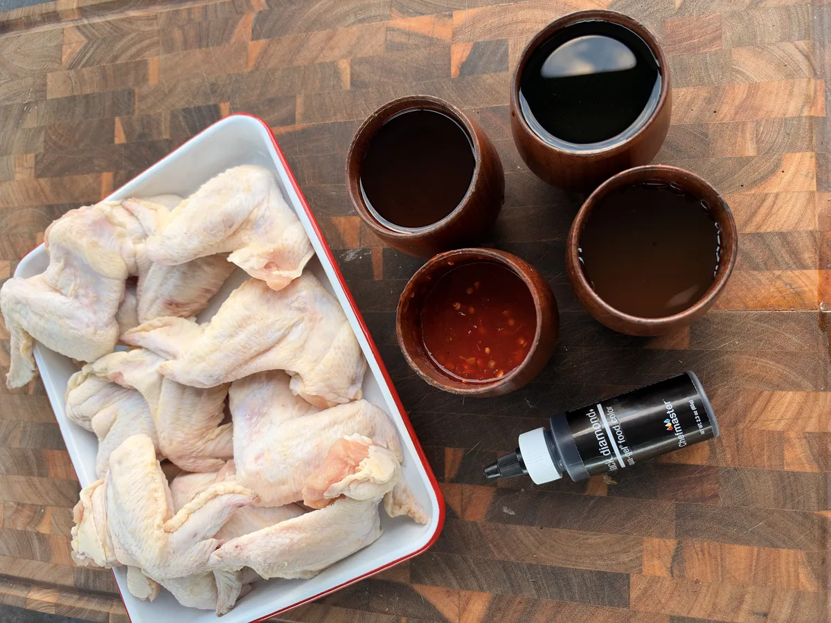Raw jumbo chicken wings in white dish with black marinade ingredients: soy sauce, honey, chili paste, and black food coloring on cutting board