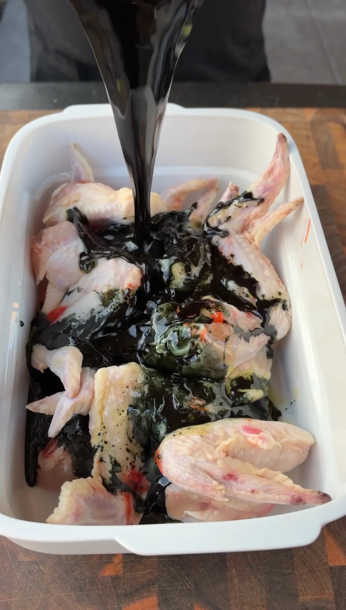 Black soy-honey-chili marinade being poured over raw jumbo chicken wings in a white container