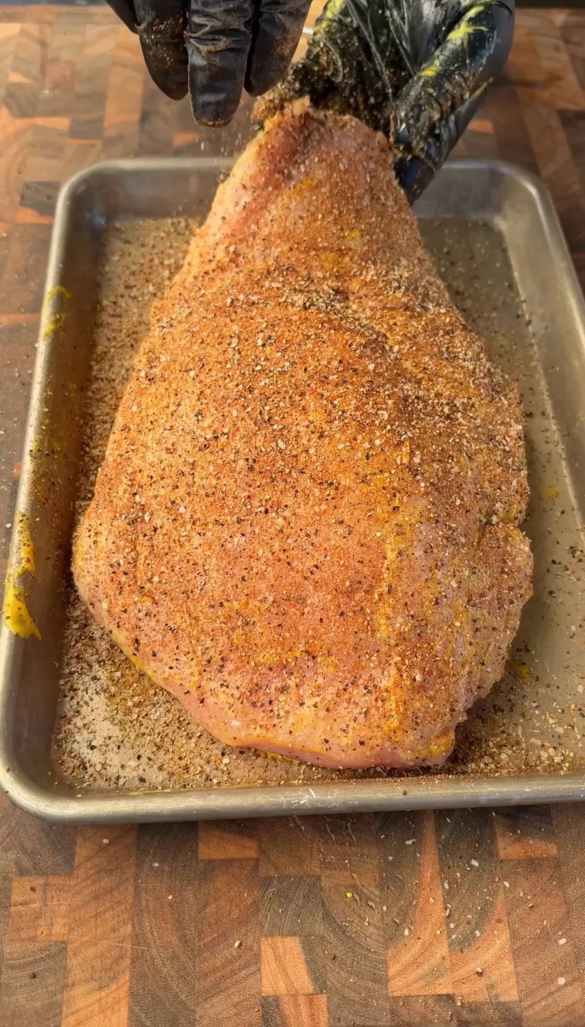 Hands in black gloves applying Texas-style salt, pepper, garlic, and paprika rub over yellow mustard-coated boneless turkey breast on sheet pan