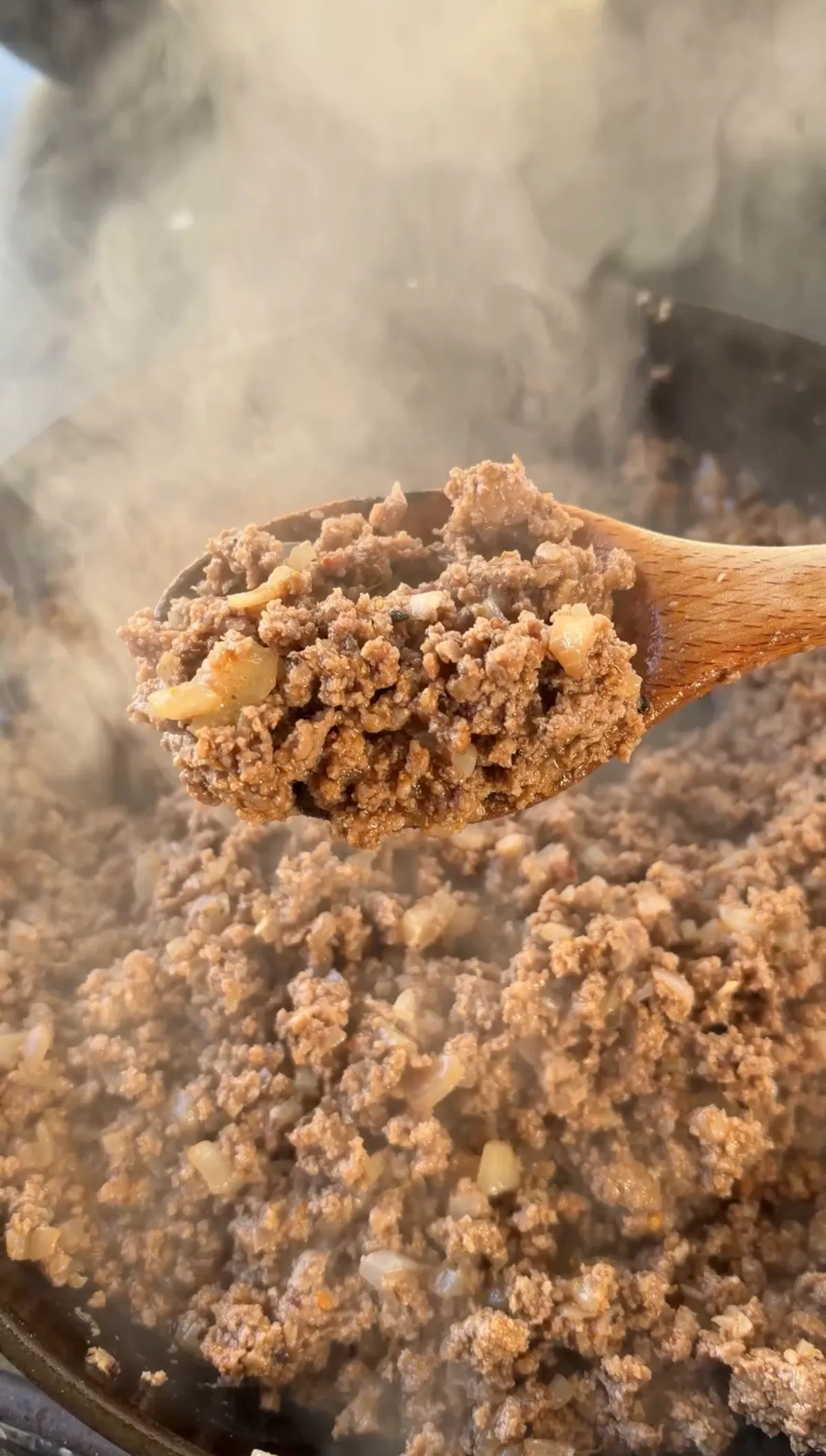 Seasoned 85/15 ground beef browned with minced garlic and onions in a steaming skillet, lifted on a wooden spoon