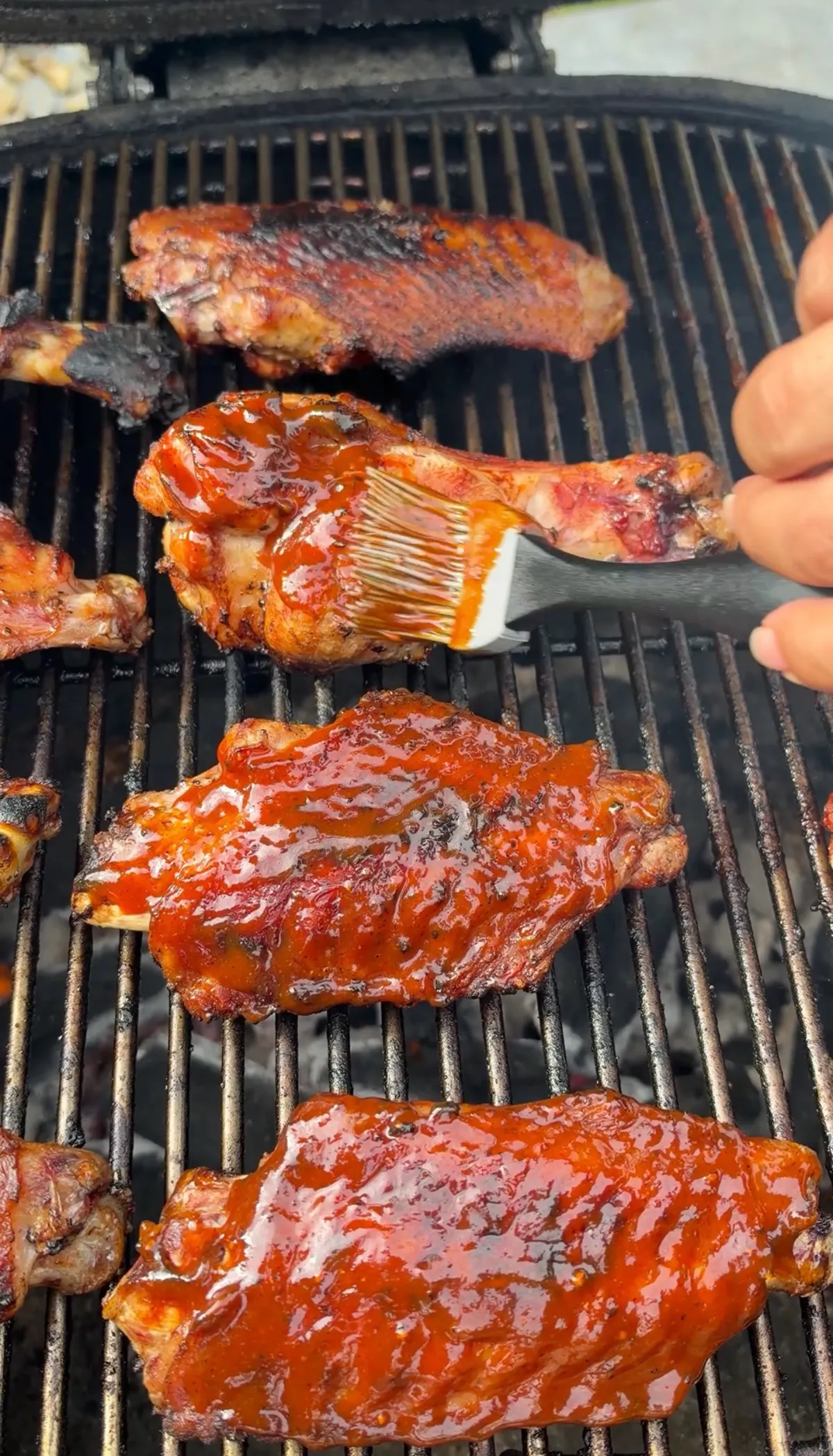 Hand brushing BBQ sauce onto grilled turkey wing drums and flats on hot grill grates during glaze step
