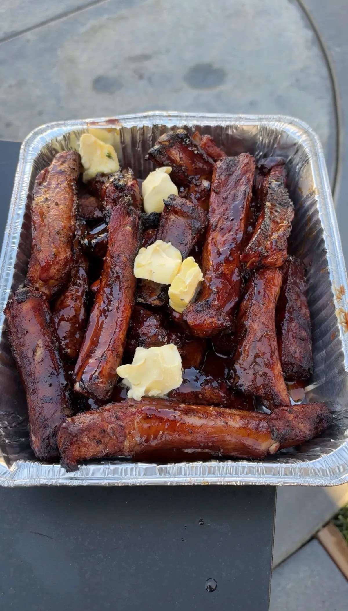 Sticky smoked barbecue party ribs piled in a foil tray with pats of unsalted butter melting over the glossy sauce