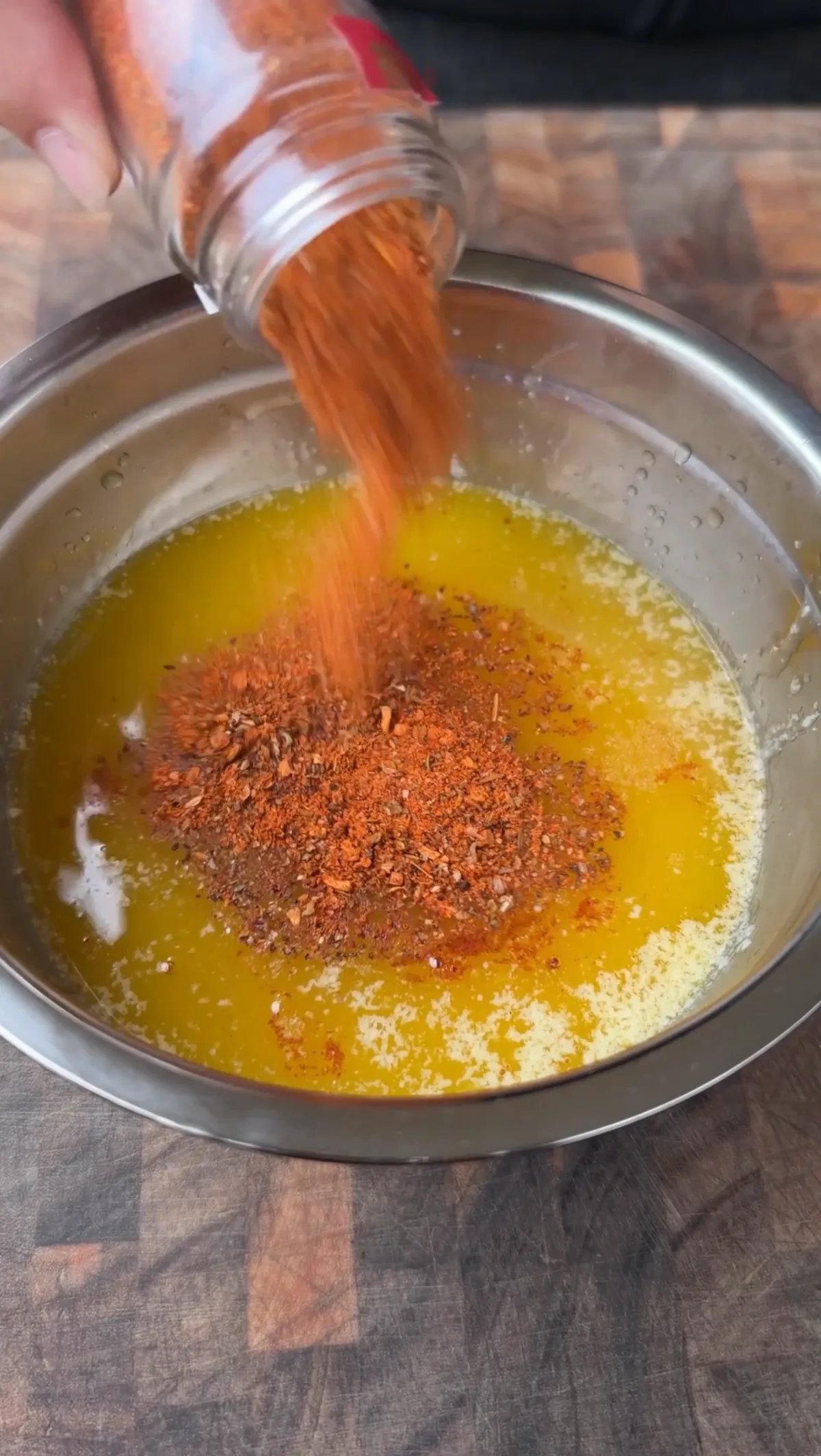 Cajun seasoning being poured from a jar into melted butter and chicken broth in a stainless steel bowl