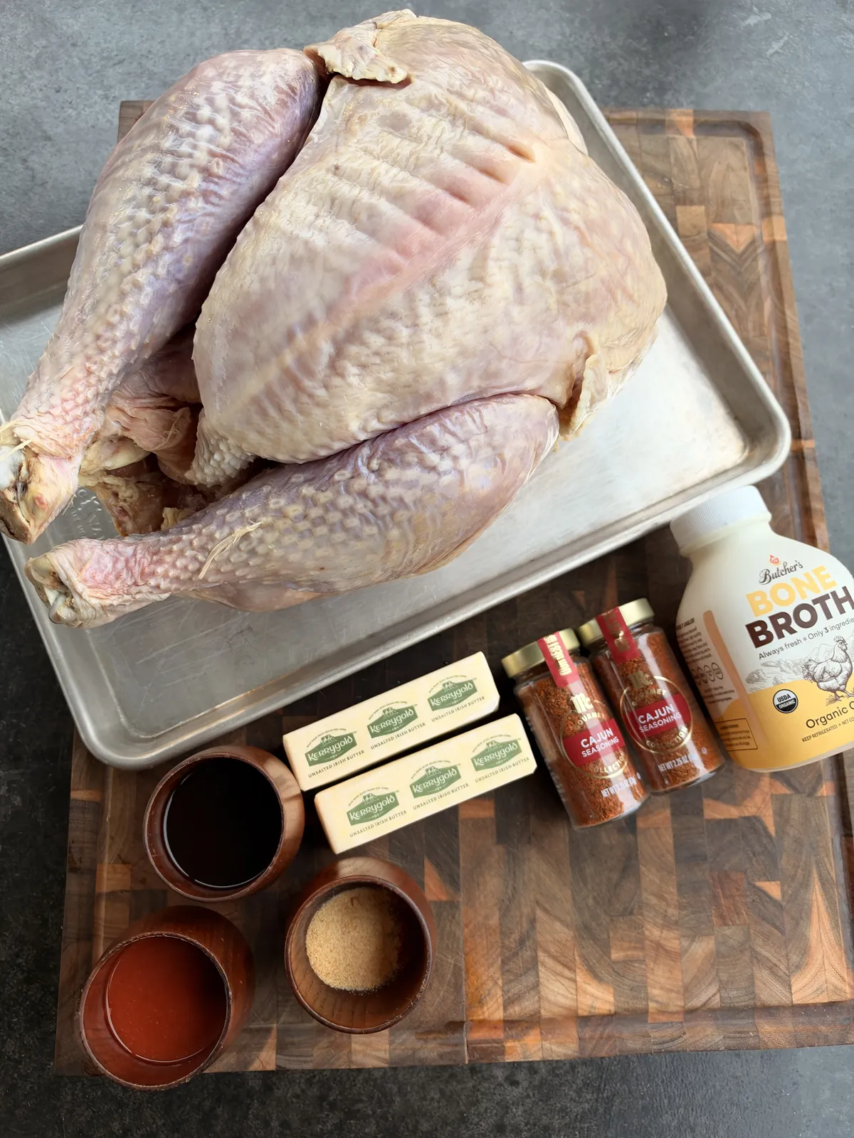 Raw whole turkey with Kerrygold butter, Cajun seasoning, bone broth, Worcestershire sauce, hot sauce, garlic powder, and smoked paprika ingredients laid out on a wooden cutting board