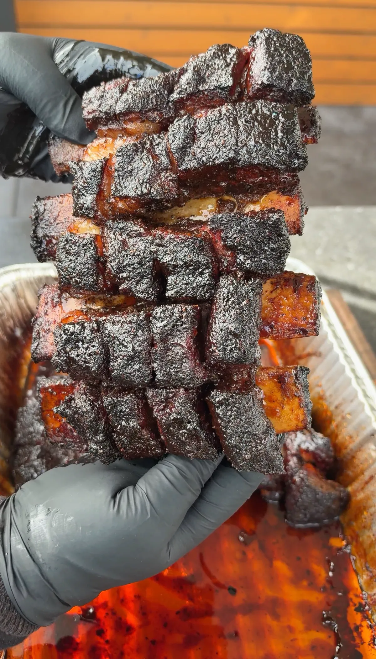 Hands in black gloves lifting a rack of deeply charred, glazed beef short rib burnt ends from a foil pan