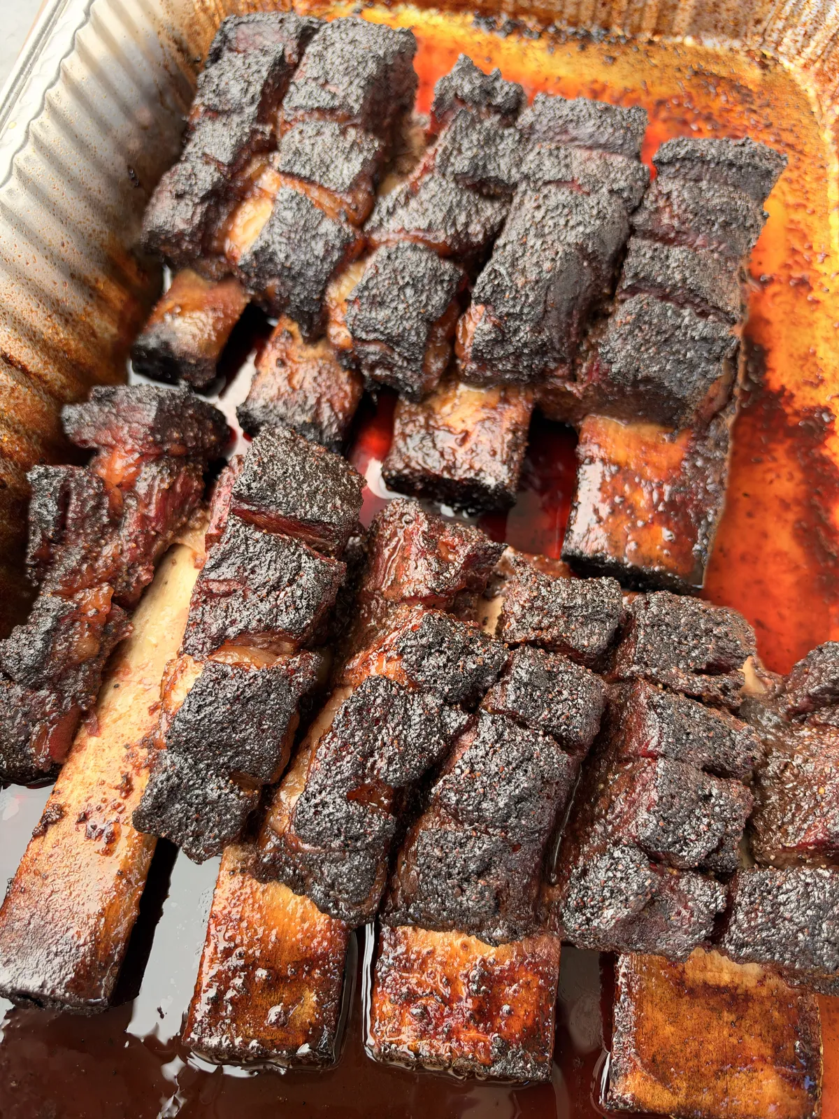 Smoked beef rib pieces with dark spice bark resting in rendered drippings inside an aluminum foil pan