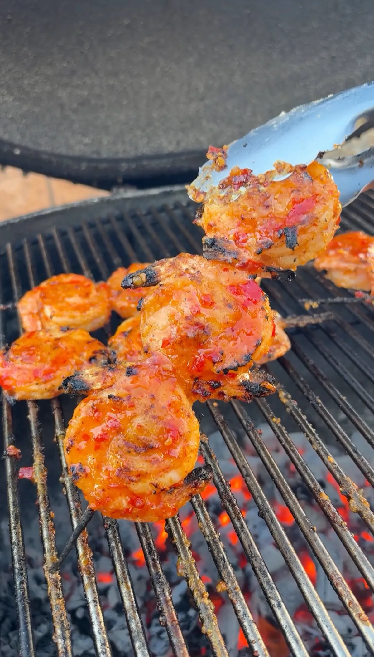 Tongs flipping sticky firecracker-glazed shrimp skewers over glowing charcoal grill grates