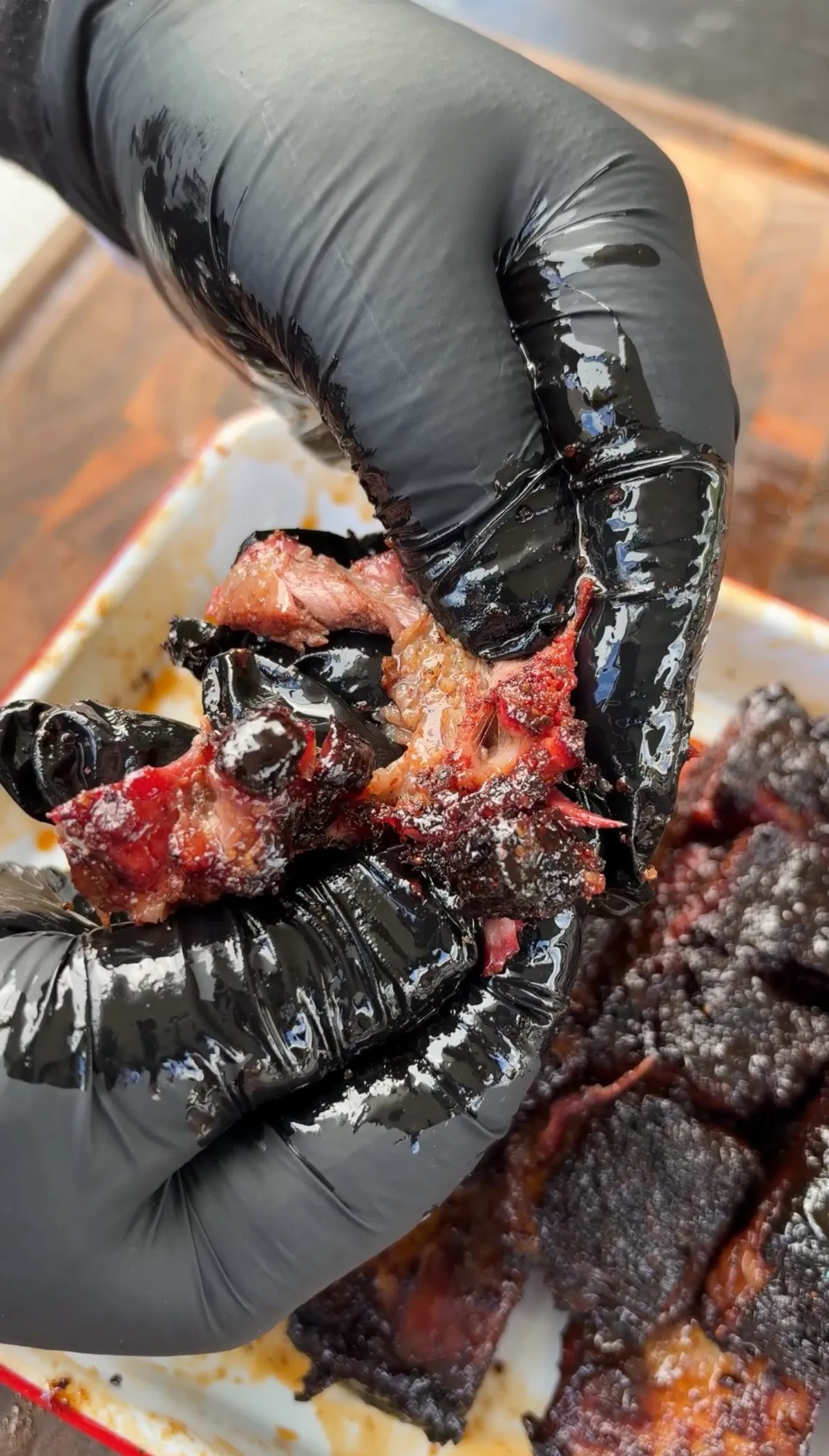 Black-gloved hands breaking apart smoky, lacquered beef short rib burnt ends revealing tender pink interior over a tray