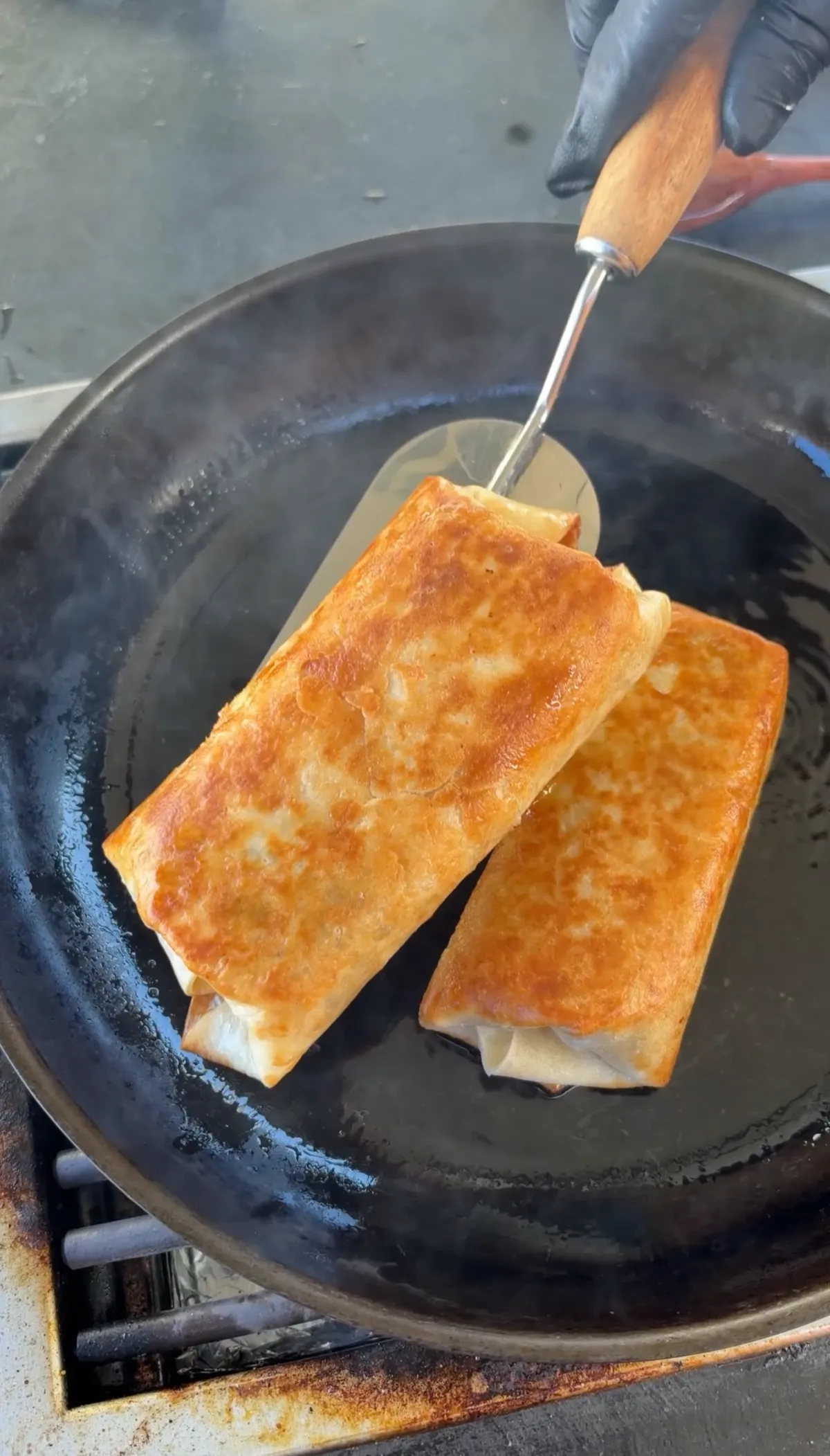 Two flour tortilla cheeseburger burritos frying golden brown in oil in a cast iron skillet, being lifted with a spatula