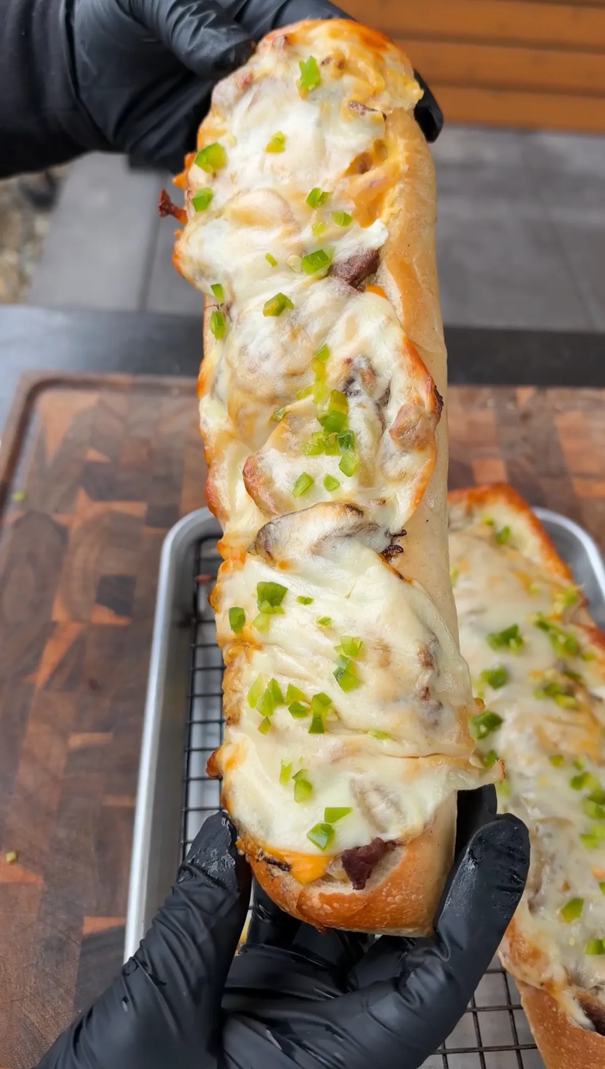 Hands in black gloves lifting a cheesesteak stuffed baguette with melted provolone and diced jalapeños over a wire rack pan