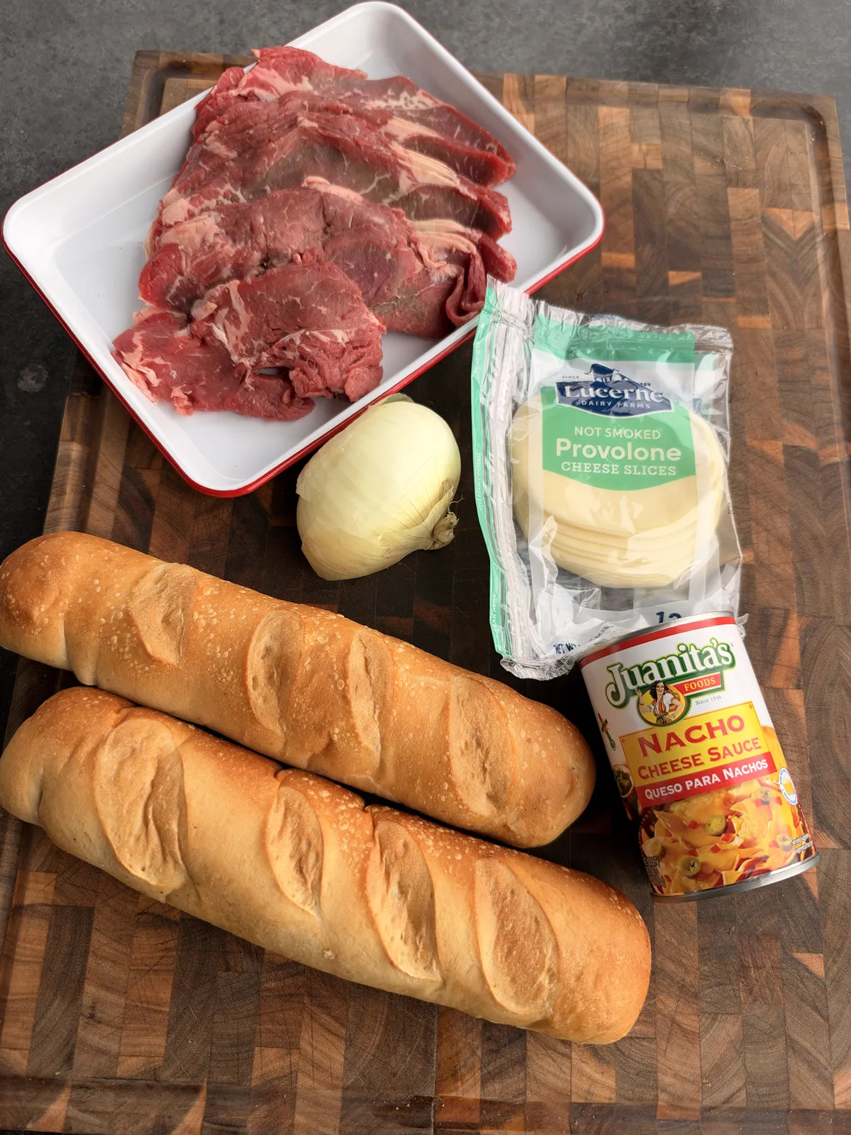 Cheesesteak bread boat ingredients: ribeye steak, sweet onion, provolone slices, nacho cheese sauce, and French baguettes on cutting board