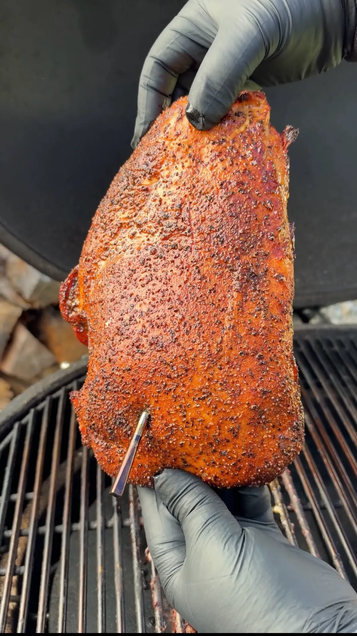 Hands in black gloves holding a seasoned boneless turkey breast with salt, pepper, and garlic rub over smoker grates
