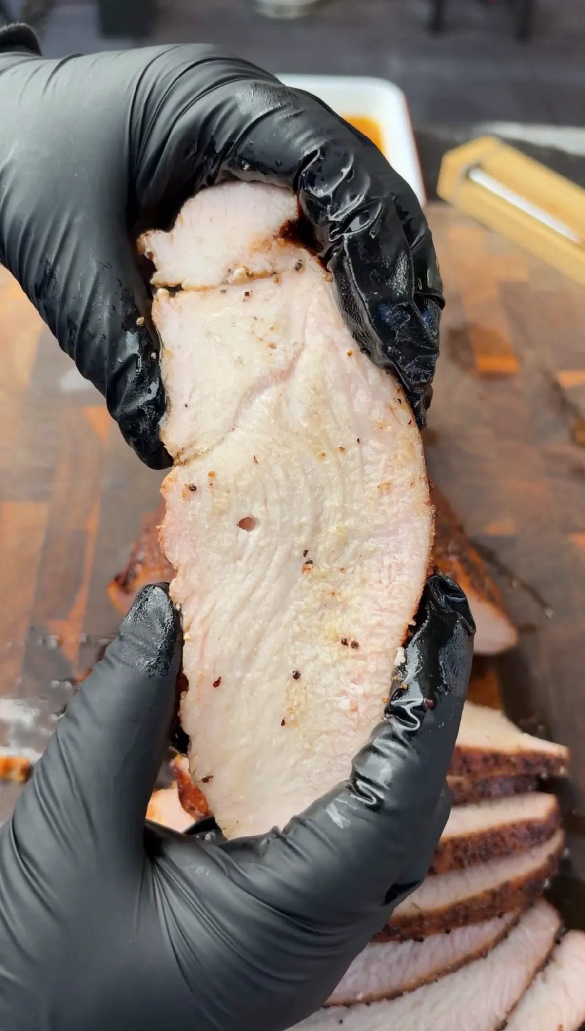 Sliced cherrywood smoked turkey breast held in black gloves showing juicy interior with salt and pepper rub crust