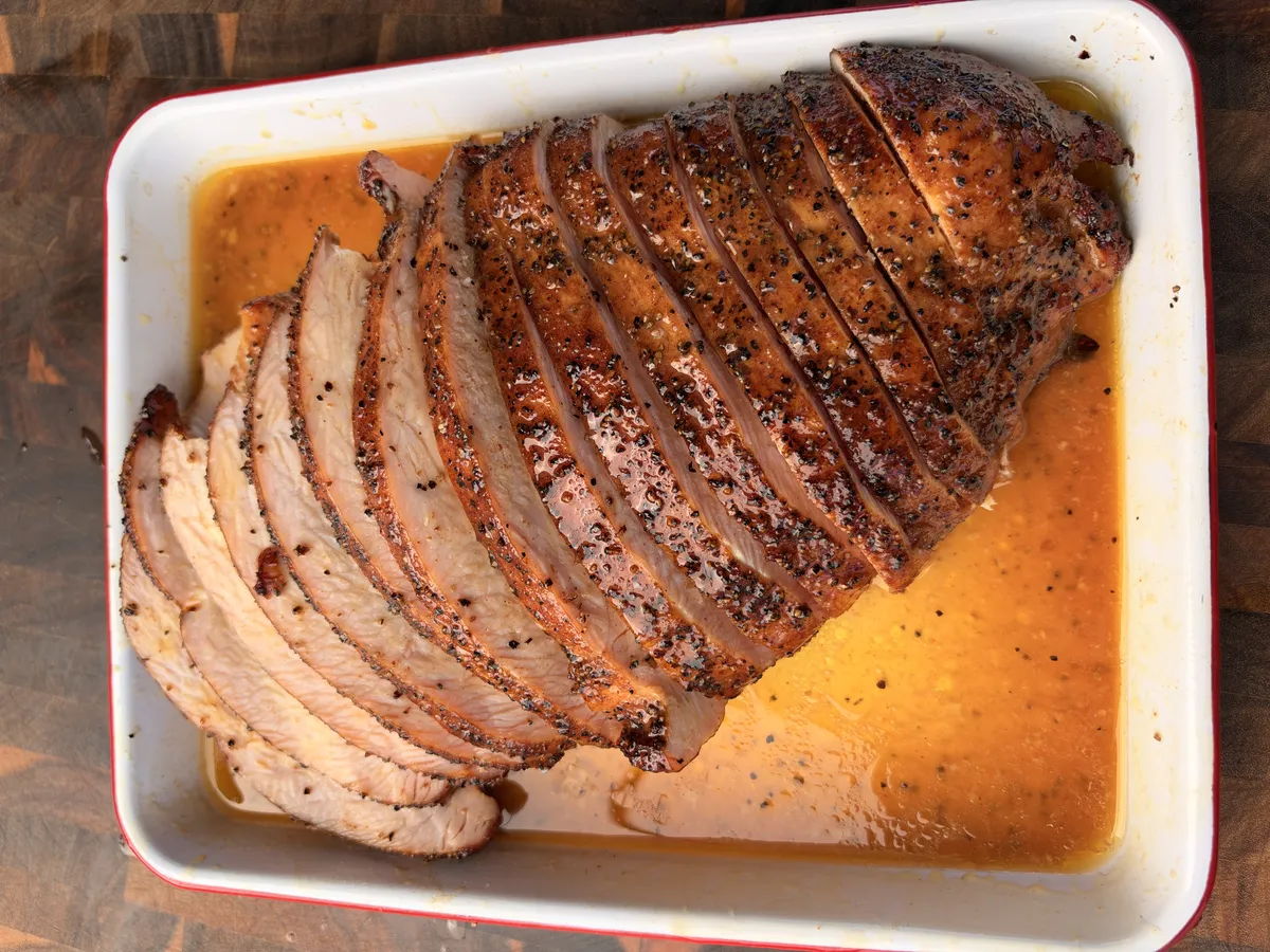 Sliced cherrywood smoked turkey breast with salt and pepper crust resting in melted honey butter juices in a white tray
