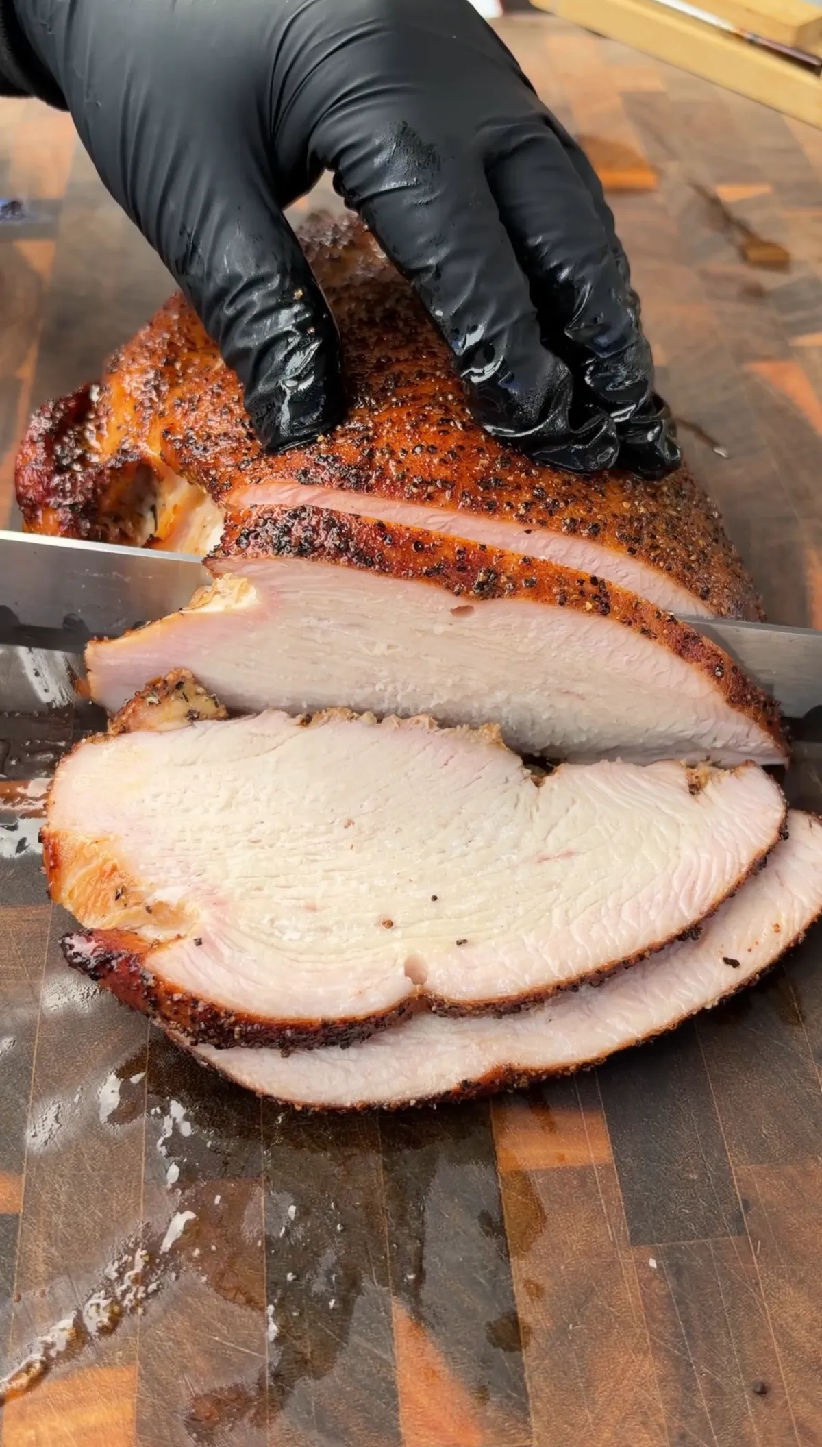 Gloved hands slicing cherrywood smoked turkey breast with salt, pepper, and garlic rub on a wooden cutting board