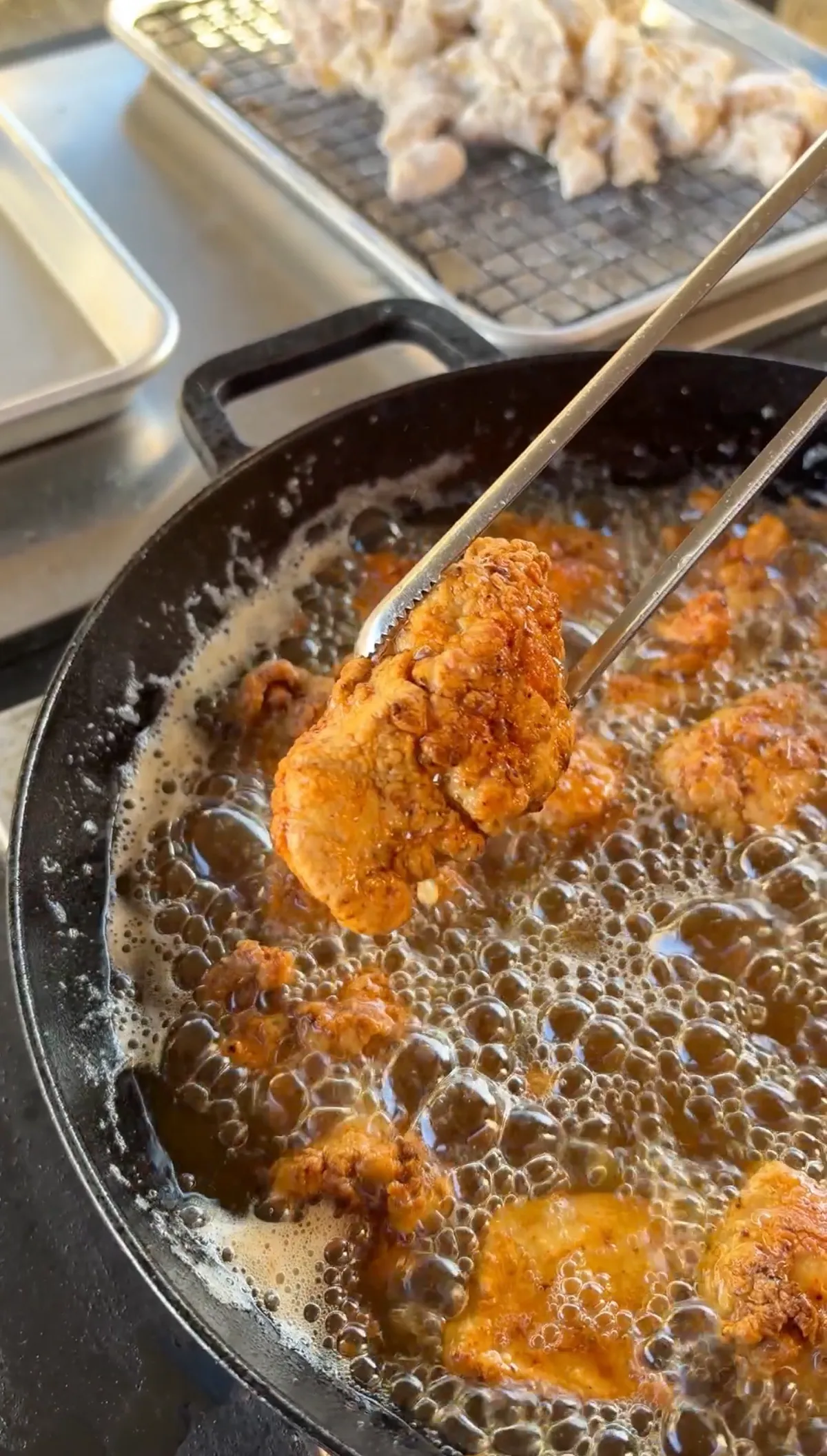 Tongs lifting a golden-brown fried chicken thigh nugget from bubbling hot oil in a cast iron skillet