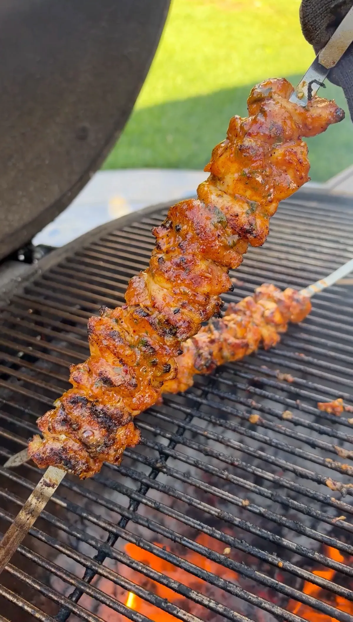 Grilled chicken thigh skewer being lifted off charcoal grill with tongs, showing char and cowboy butter glaze