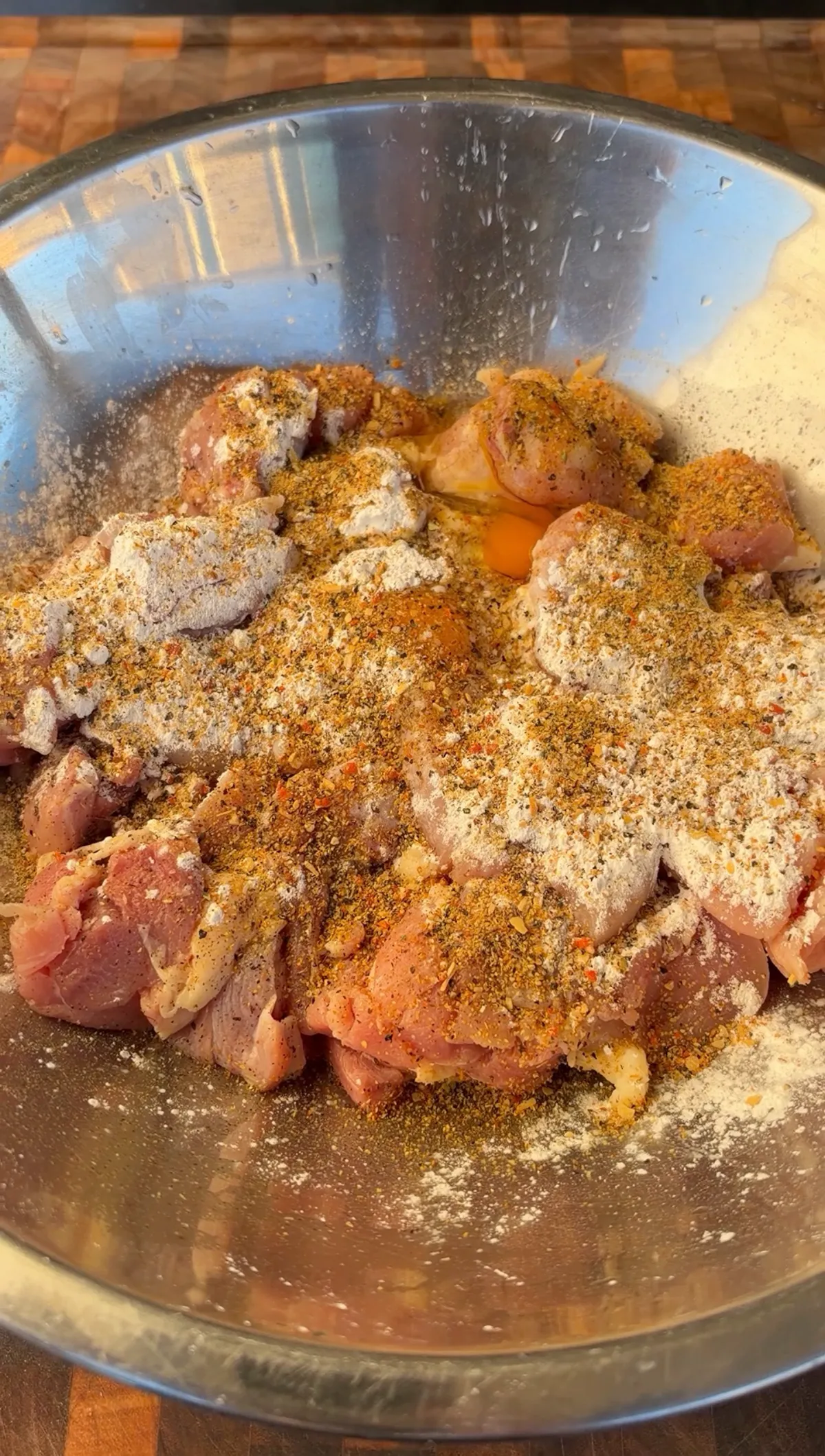 Raw chicken thigh pieces in a stainless steel bowl topped with lemon pepper seasoning, cornstarch, and cracked eggs before mixing