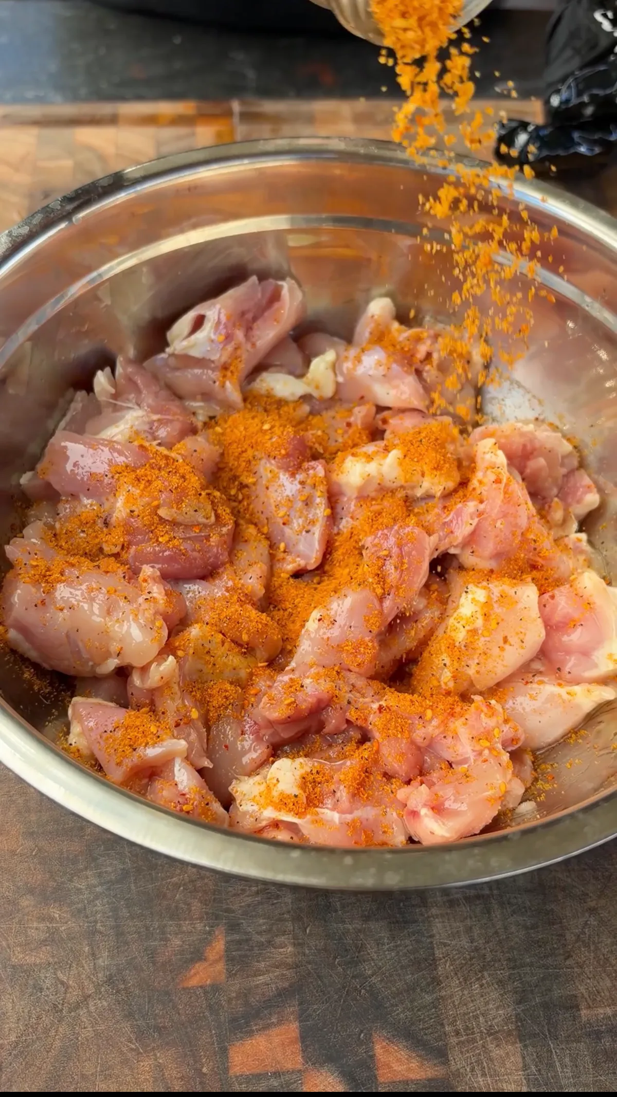 Poultry seasoning being poured over raw chicken thigh pieces in a stainless steel mixing bowl