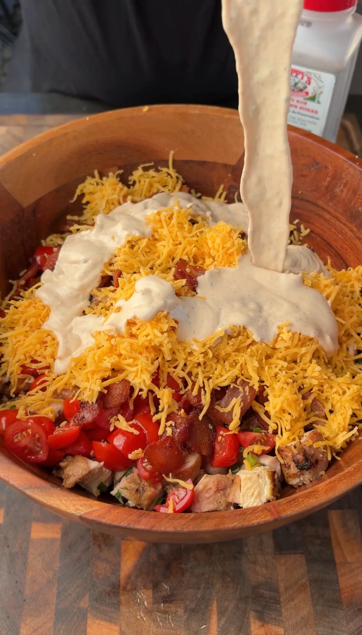 Creamy chipotle ranch dressing being poured over grilled chicken, crispy bacon, cherry tomatoes, and cheddar cheese in a wooden bowl