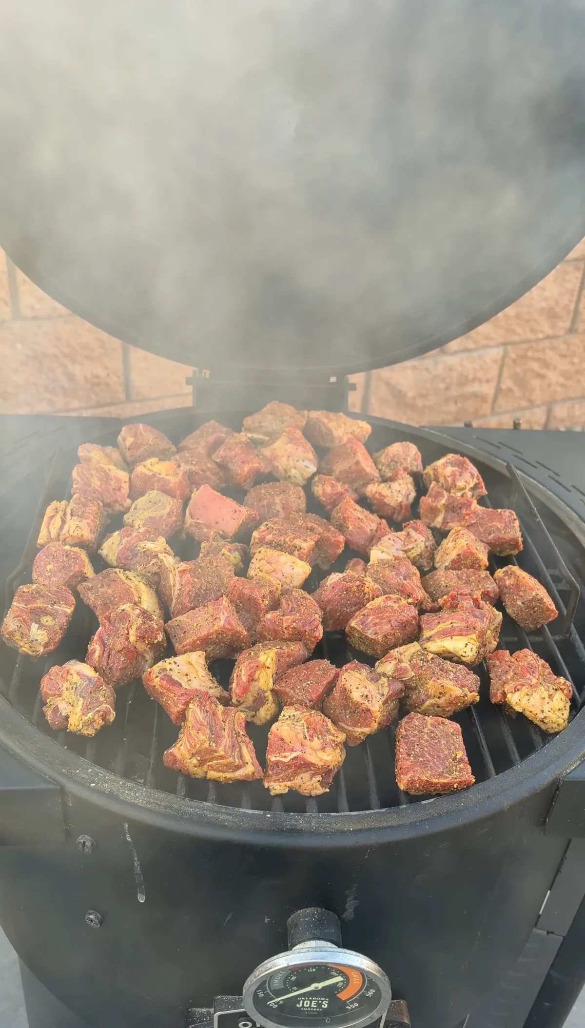 Seasoned chuck roast cubes smoking on an Oklahoma Joe's smoker grates with cherry wood smoke rising